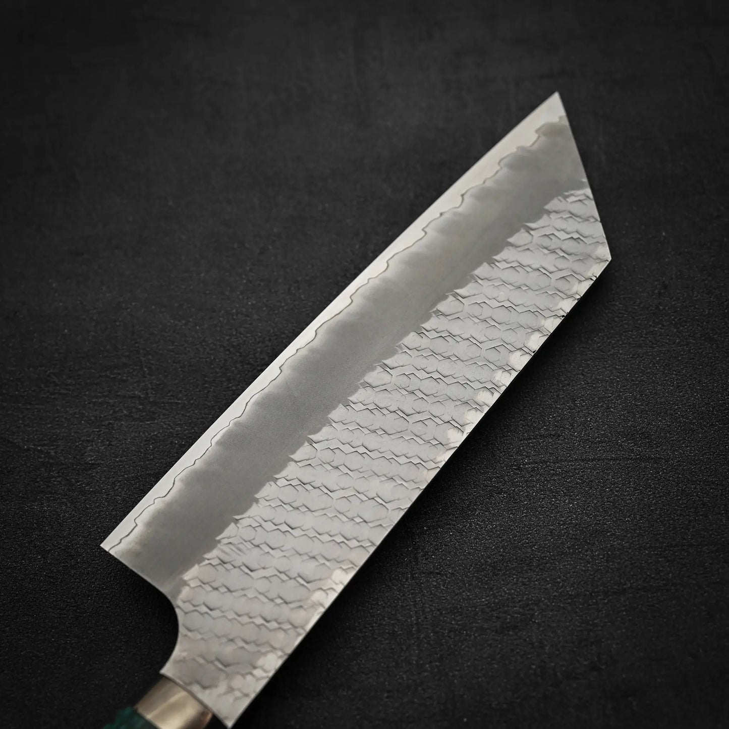 A close-up of the Nigara Hamono Nigara tsuchime SG2 kiritsuke nakiri 180mm knife, featuring a textured hammered SG2 stainless steel blade with a visible pattern, set against a dark background—ideal for precise vegetable prep.