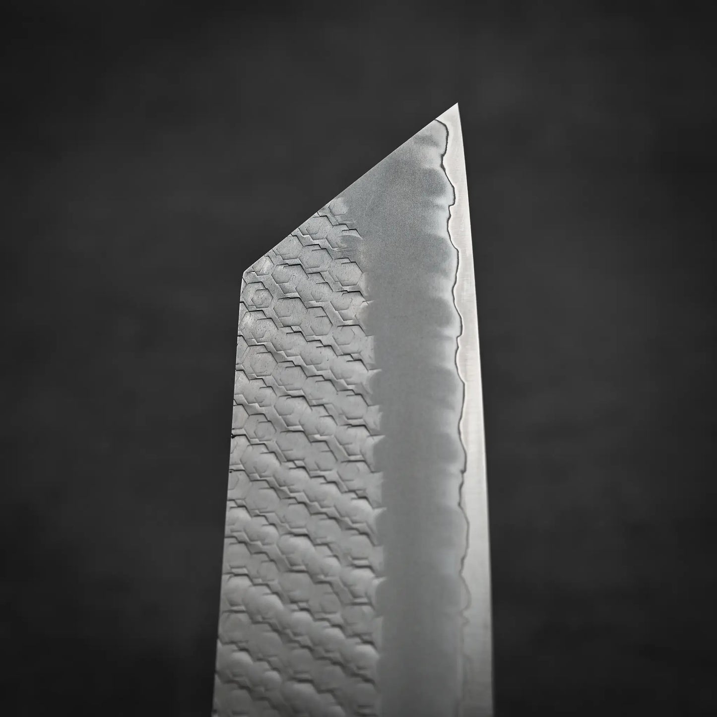 A close-up of the Nigara Hamono Nigara tsuchime SG2 kiritsuke nakiri 180mm shows its patterned SG2 stainless steel blade, sharp edge, and wavy lines, set against a dark, blurred background.