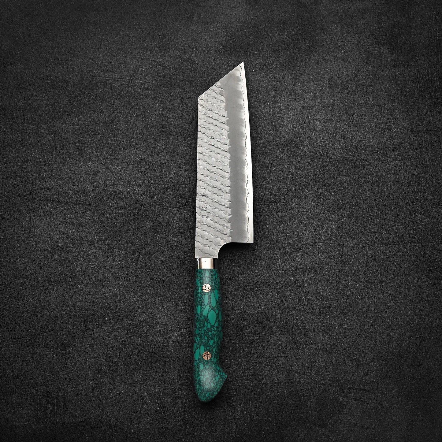 The Nigara Hamono Nigara tsuchime SG2 kiritsuke nakiri 180mm, featuring a green handle and a textured silver SG2 stainless steel blade, rests on a dark surface—ideal for precise vegetable prep.