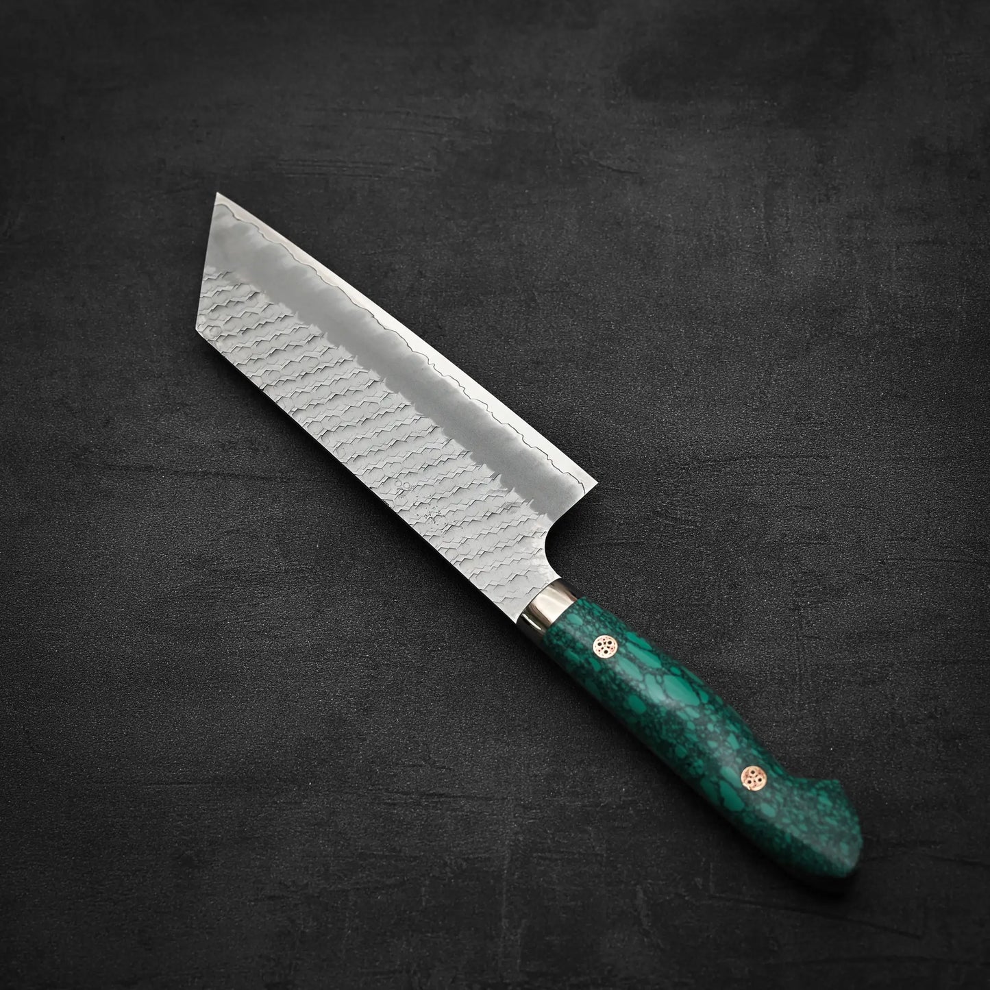 The Nigara Hamono Nigara tsuchime SG2 kiritsuke nakiri 180mm, featuring a patterned SG2 stainless steel blade and green marbled handle, rests on a dark surface—ideal for precision vegetable prep.