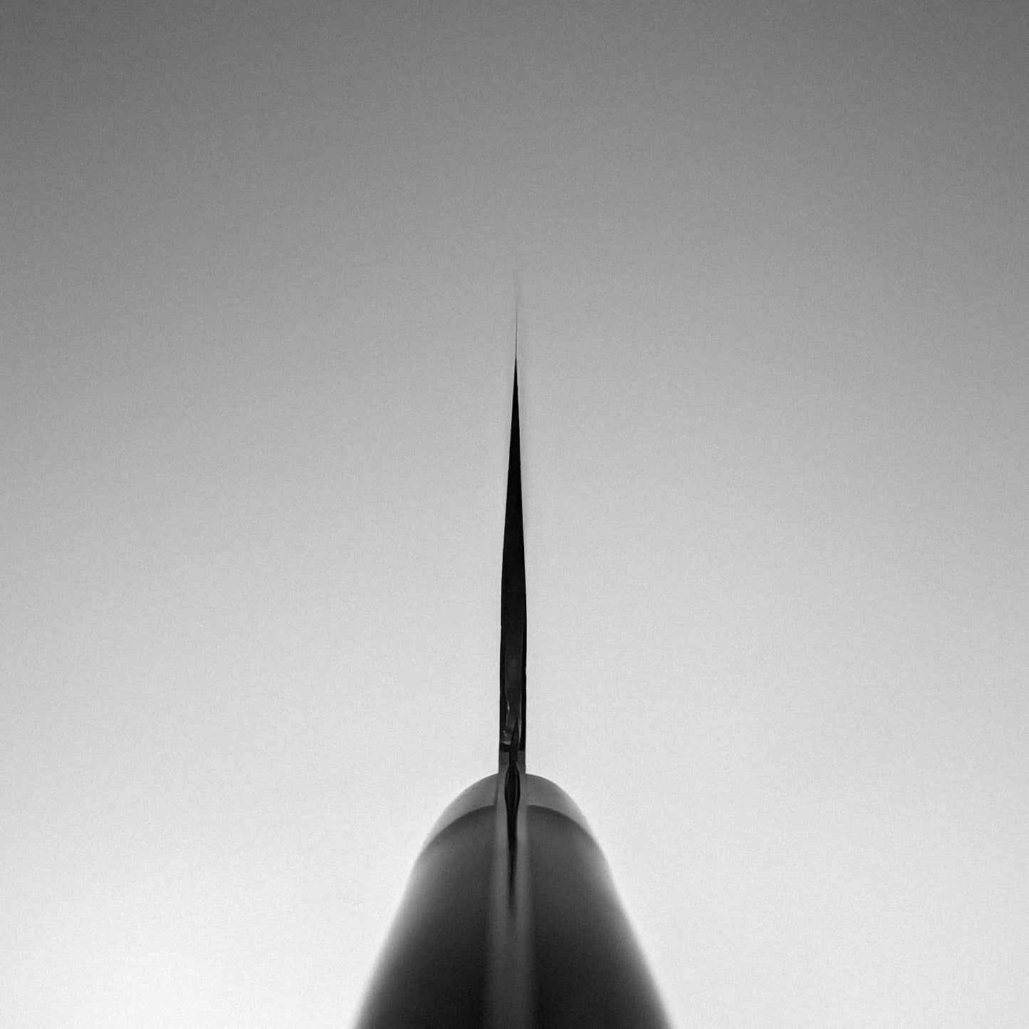 A close-up, black-and-white photo captures the tip of the Nigara Hamono Nigara tsuchime SG2 kiritsuke nakiri 180mm blade, crafted from SG2 stainless steel, pointed upward and centered against a smooth gradient background.