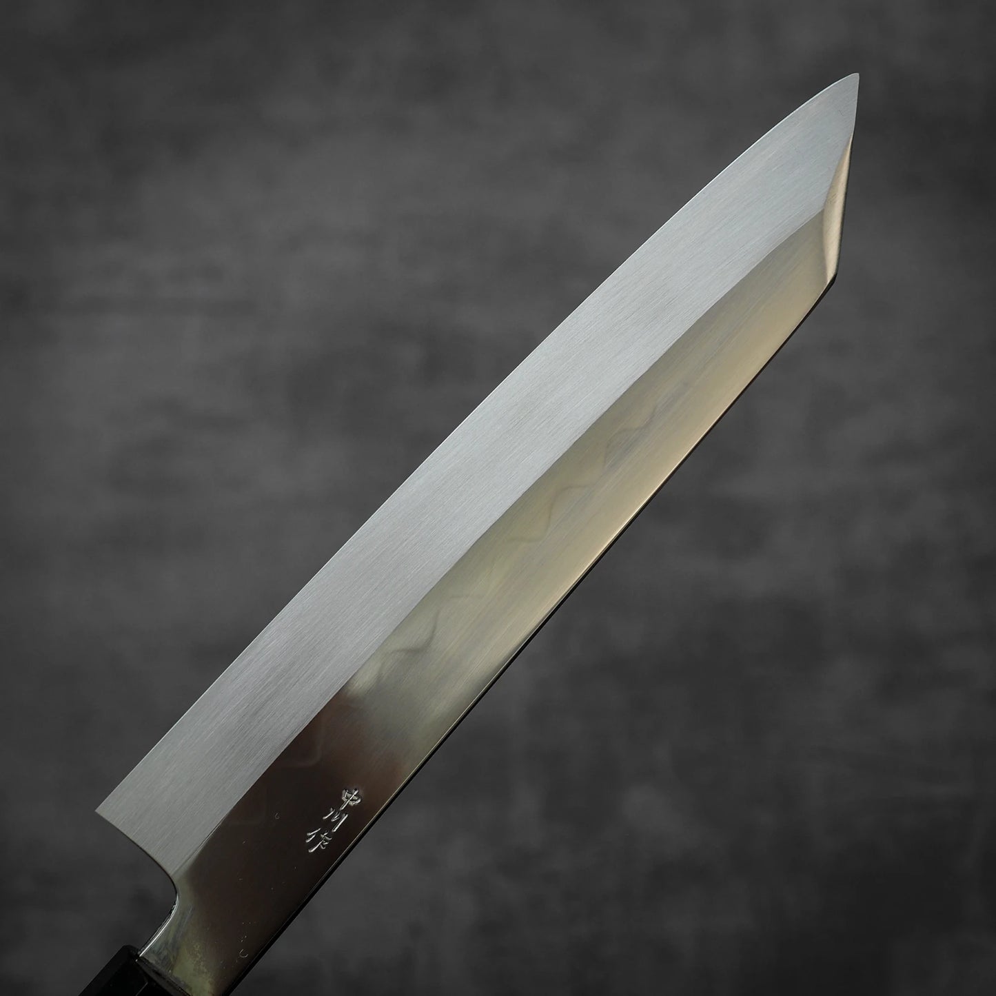 A close-up of the Satoshi Nakagawa Nakagawa shirogami#3 honyaki kiritsuke gyuto 210mm displays its straight-edged blade, pointed tip, visible steel patterns, and Japanese characters near the handle on a dark gray background.