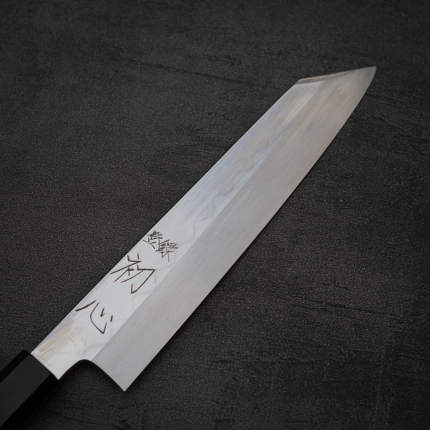 A Nakagawa shirogami#3 honyaki kiritsuke gyuto 210mm by Satoshi Nakagawa, featuring kanji-engraved blade, lies on a dark textured surface.