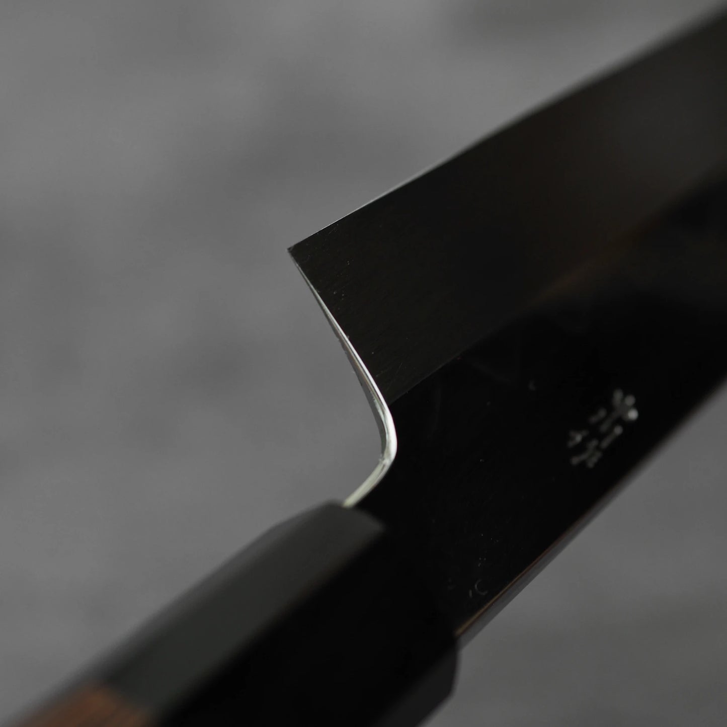 Close-up of the Satoshi Nakagawa Nakagawa shirogami#3 honyaki kiritsuke gyuto 210mm blade, highlighting its sharp edge and craftsmanship, with part of the handle visible against a blurred grey background.