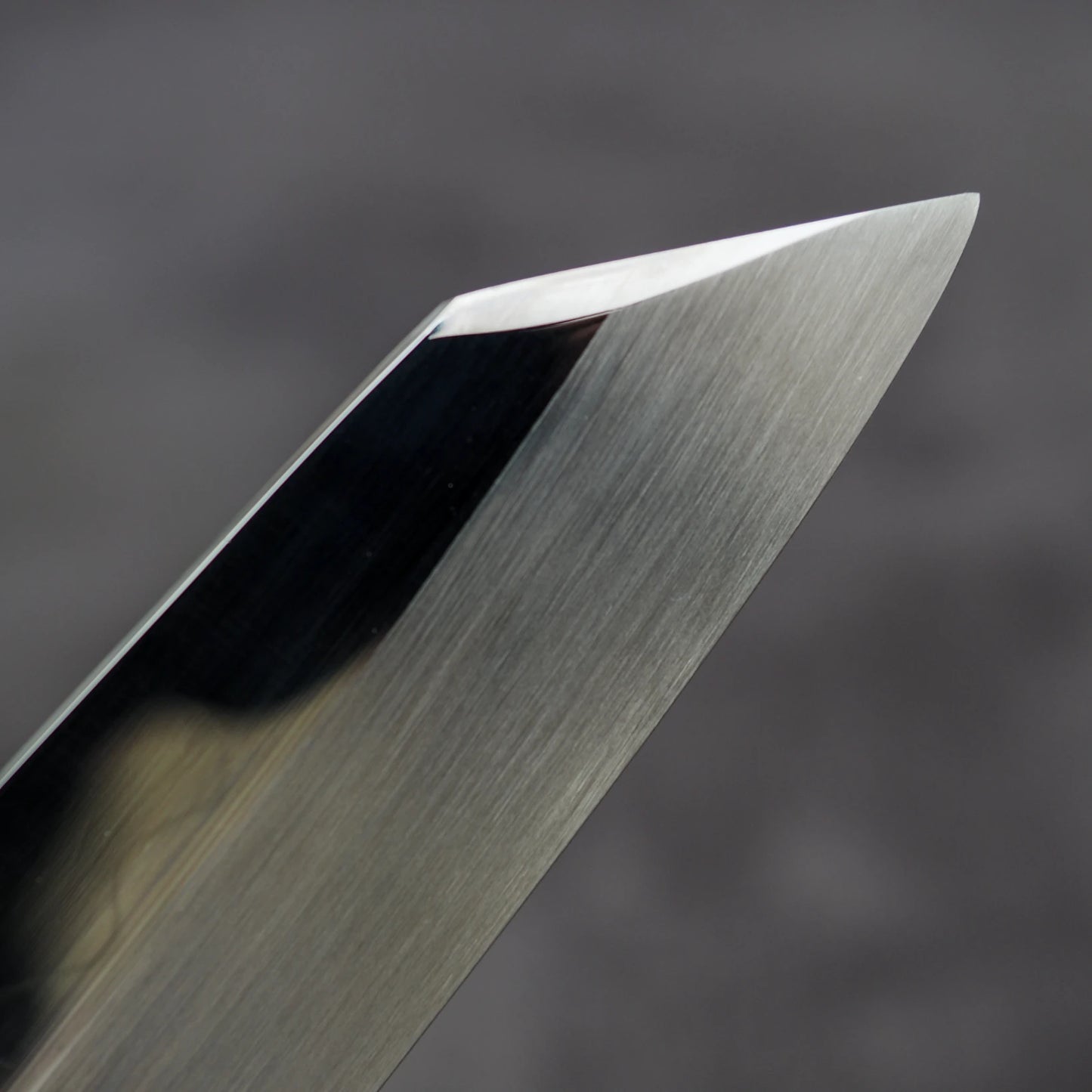 Close-up of the polished tip of the Satoshi Nakagawa Nakagawa shirogami#3 honyaki kiritsuke gyuto 210mm, displaying its Japanese craftsmanship, with clean lines and a sharp edge set against a blurred gray background.