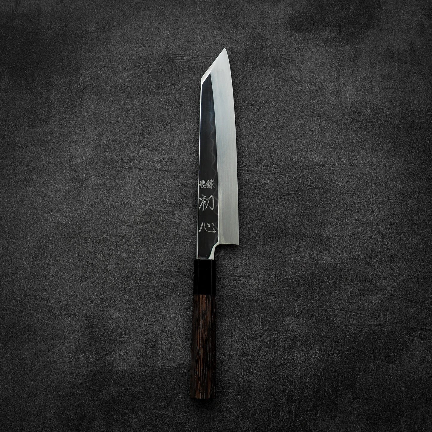 A Satoshi Nakagawa Nakagawa shirogami#3 honyaki kiritsuke gyuto 210mm Japanese kitchen knife with a dark wooden handle and engraved blade rests on a dark textured surface, photographed from above.