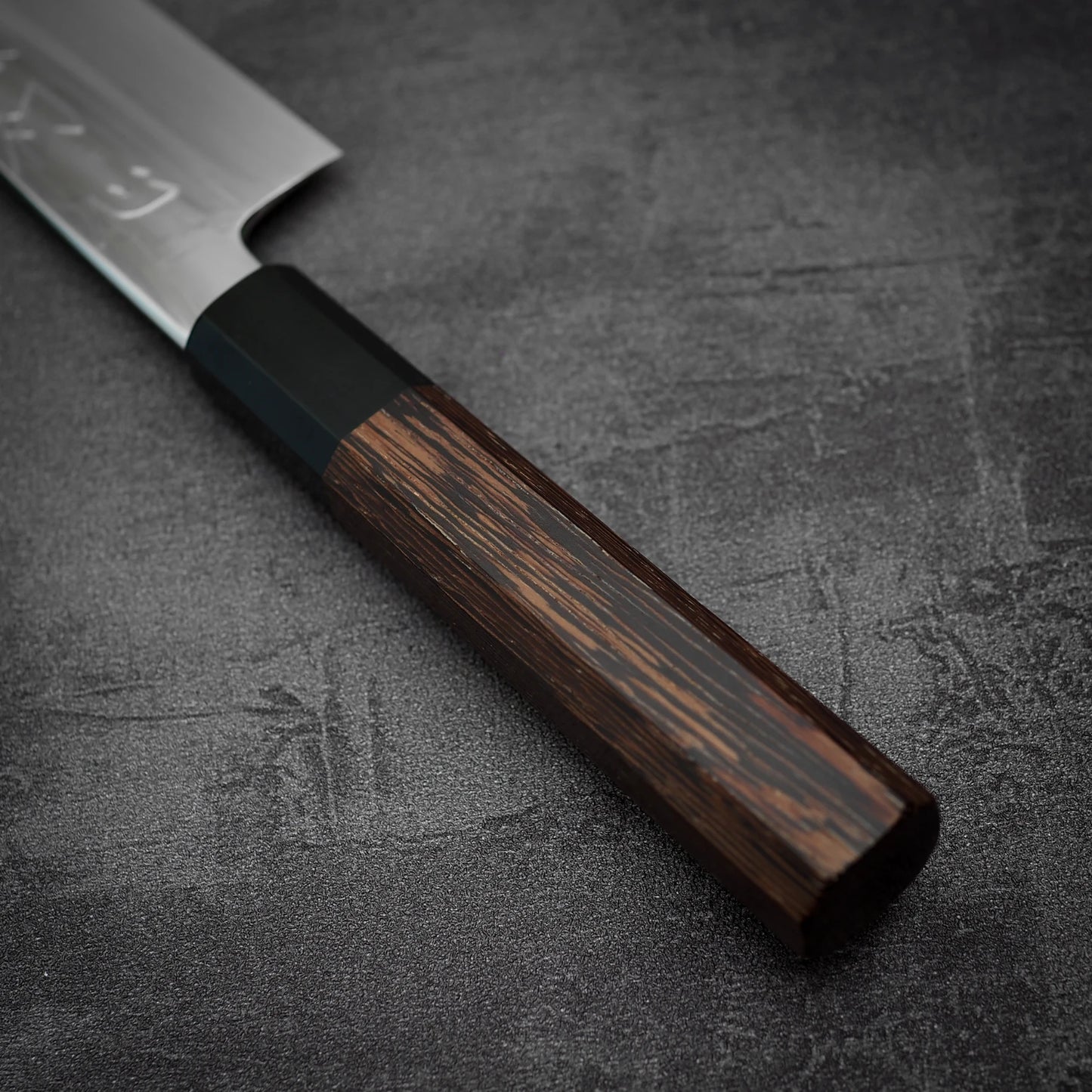 Close-up of the Satoshi Nakagawa shirogami#3 honyaki kiritsuke gyuto 210mm with a dark wooden handle and partially visible blade on a textured dark gray surface, showcasing exceptional Japanese craftsmanship.