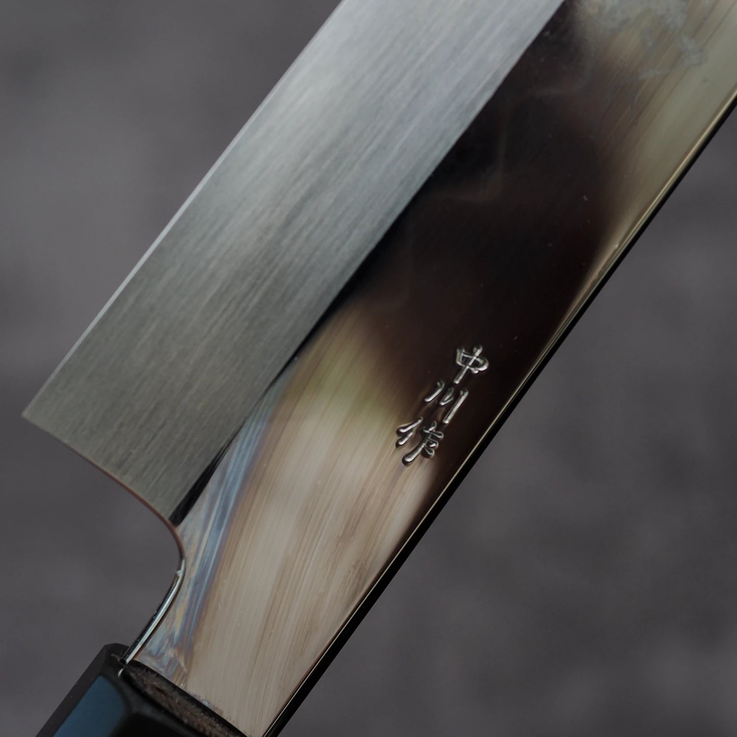 Close-up of the Nakagawa shirogami#3 honyaki kiritsuke gyuto 210mm by Satoshi Nakagawa, featuring a shiny blade and Japanese etching near the handle that highlight expert craftsmanship against a blurred gray background.