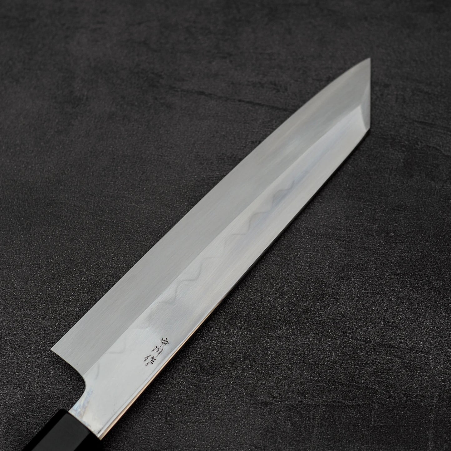 The Satoshi Nakagawa Nakagawa shirogami#3 honyaki kiritsuke gyuto 210mm features a sharp, angled blade with subtle patterns and engraved Japanese characters, partially visible on a dark textured surface.