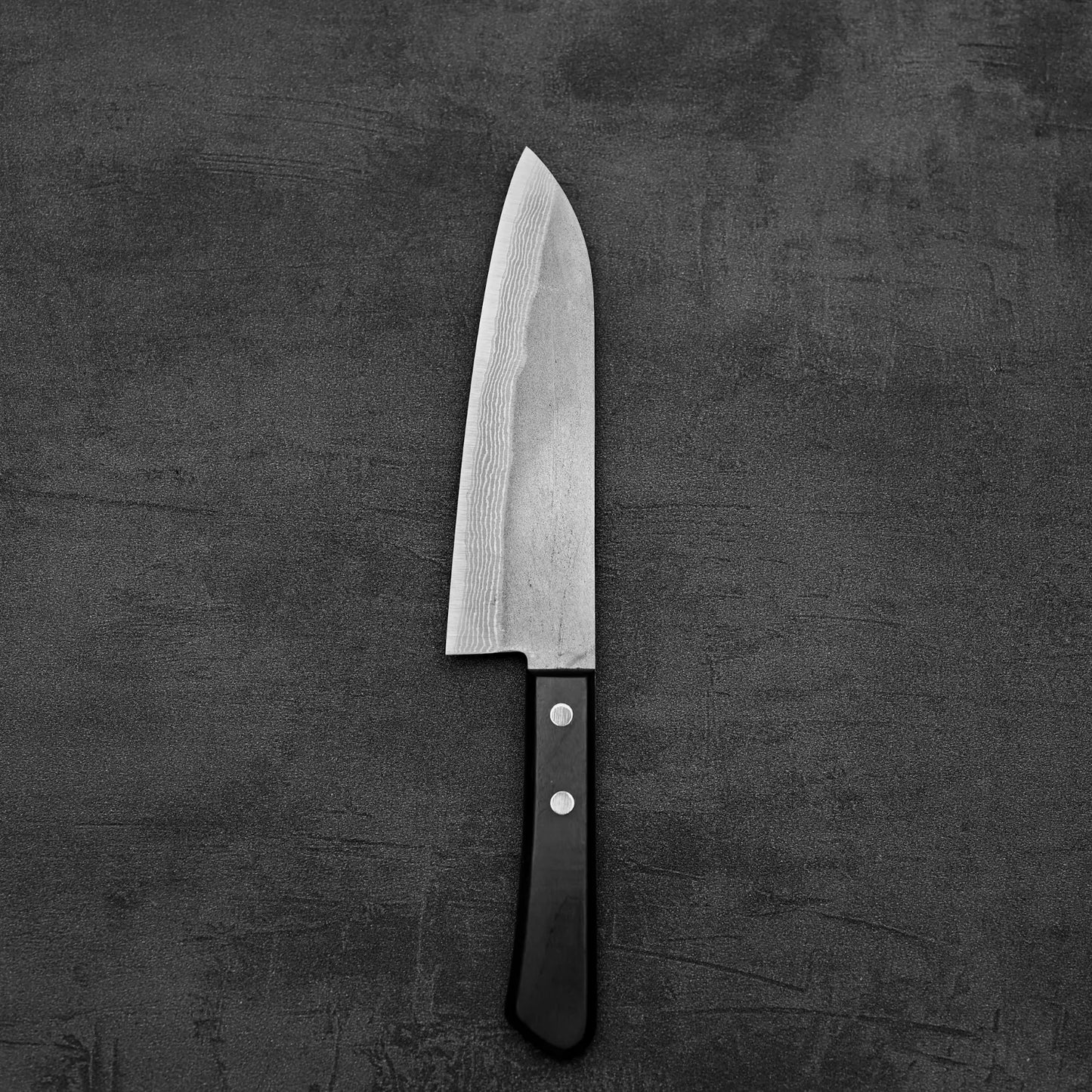 A Shigeki Tanaka nashiji damascus VG10 santoku with a black handle rests on a dark, textured surface.