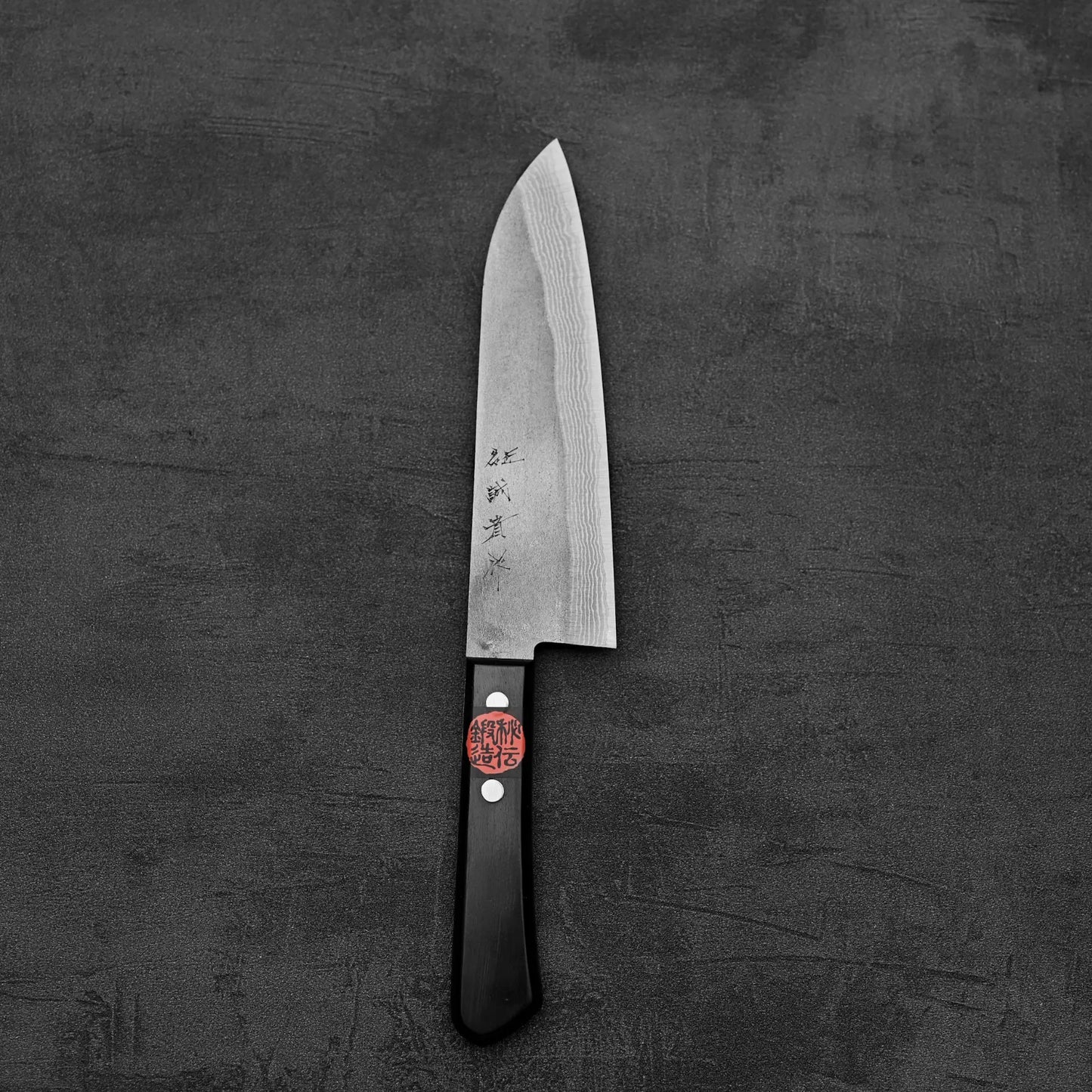 A Shigeki Tanaka nashiji damascus VG10 santoku kitchen knife with a black handle and engraved blade, bearing a red sticker, rests on a dark, textured surface.