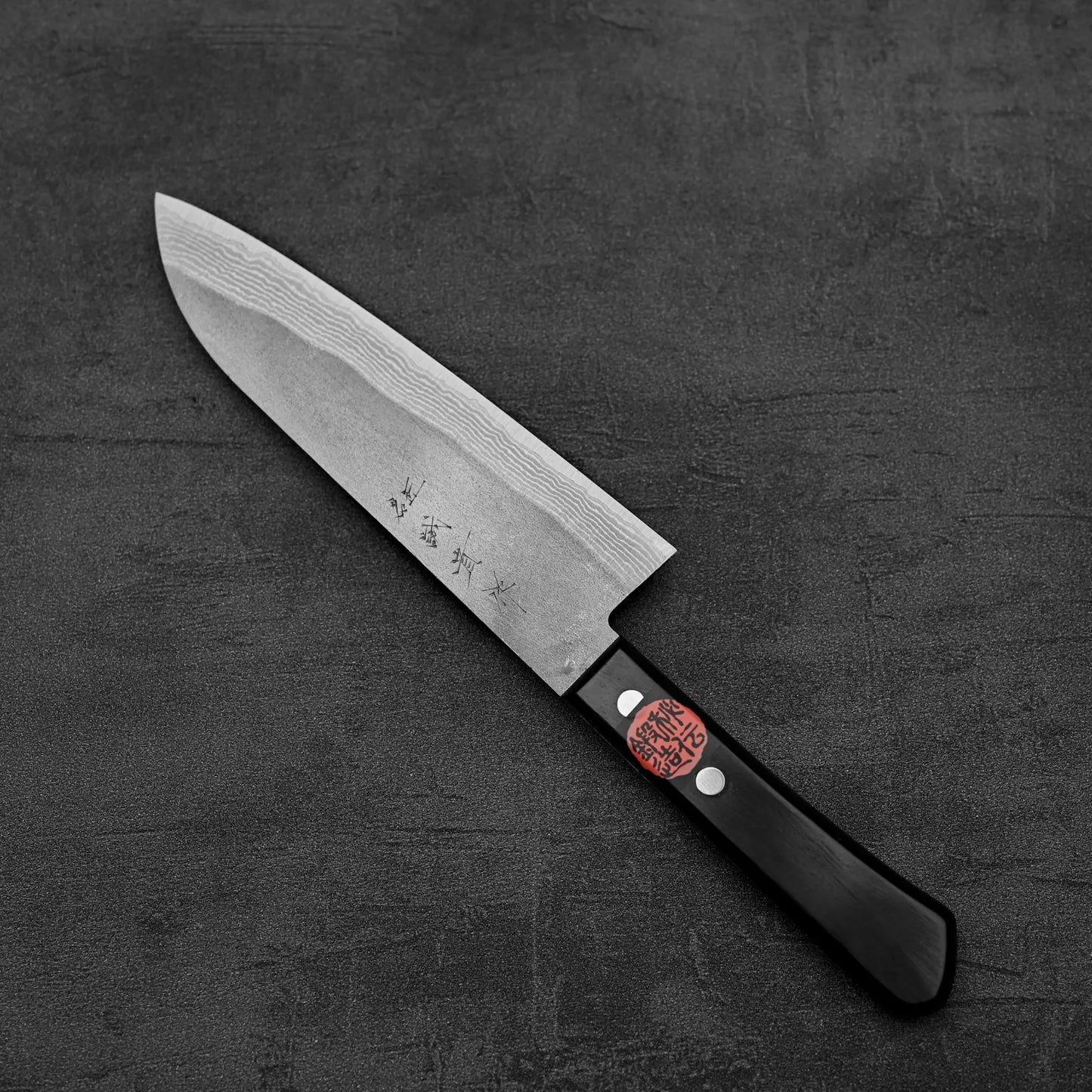 A Shigeki Tanaka nashiji damascus VG10 santoku by Shigeki Tanaka, featuring a black handle and engraved blade with a red label, rests on a dark textured surface.