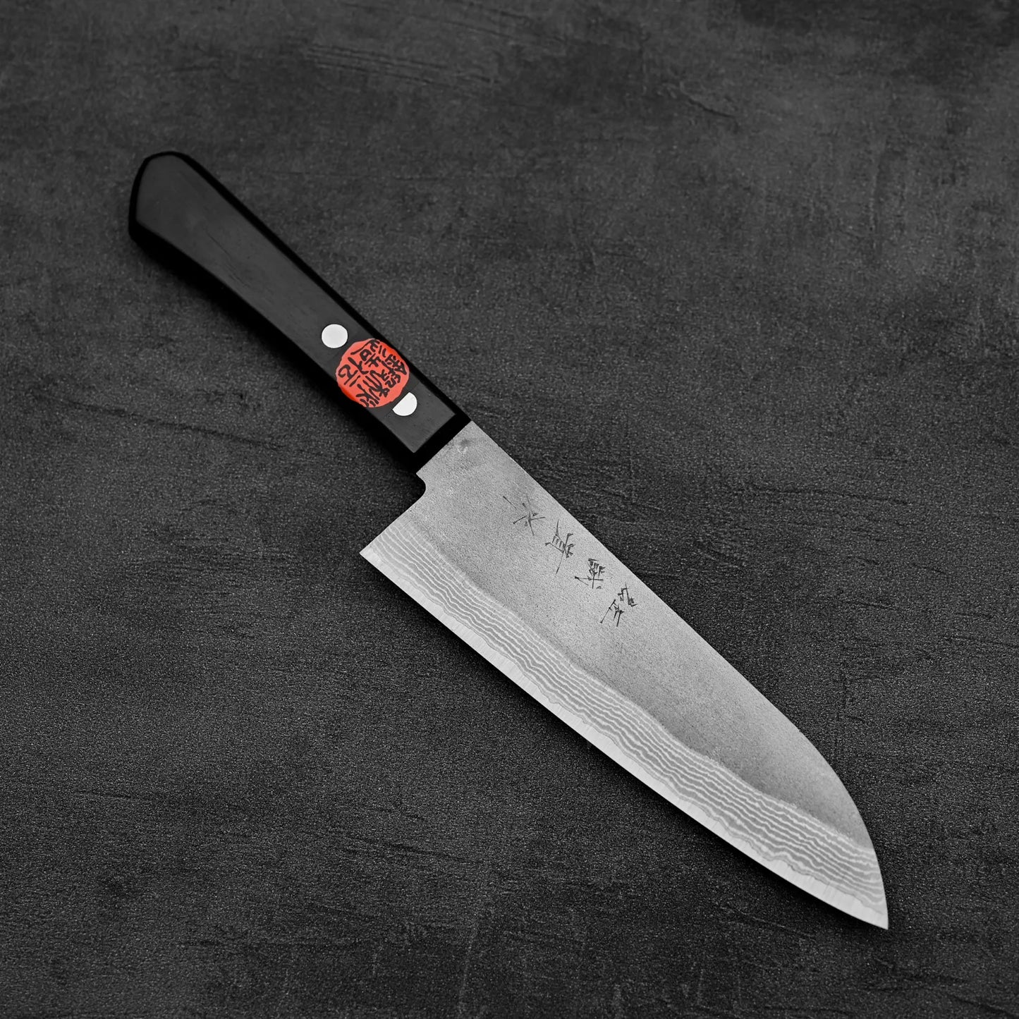 A Shigeki Tanaka nashiji damascus VG10 santoku knife with a black handle and visible damascus pattern rests on a dark textured surface. A red sticker with white and black markings lies near the handle.