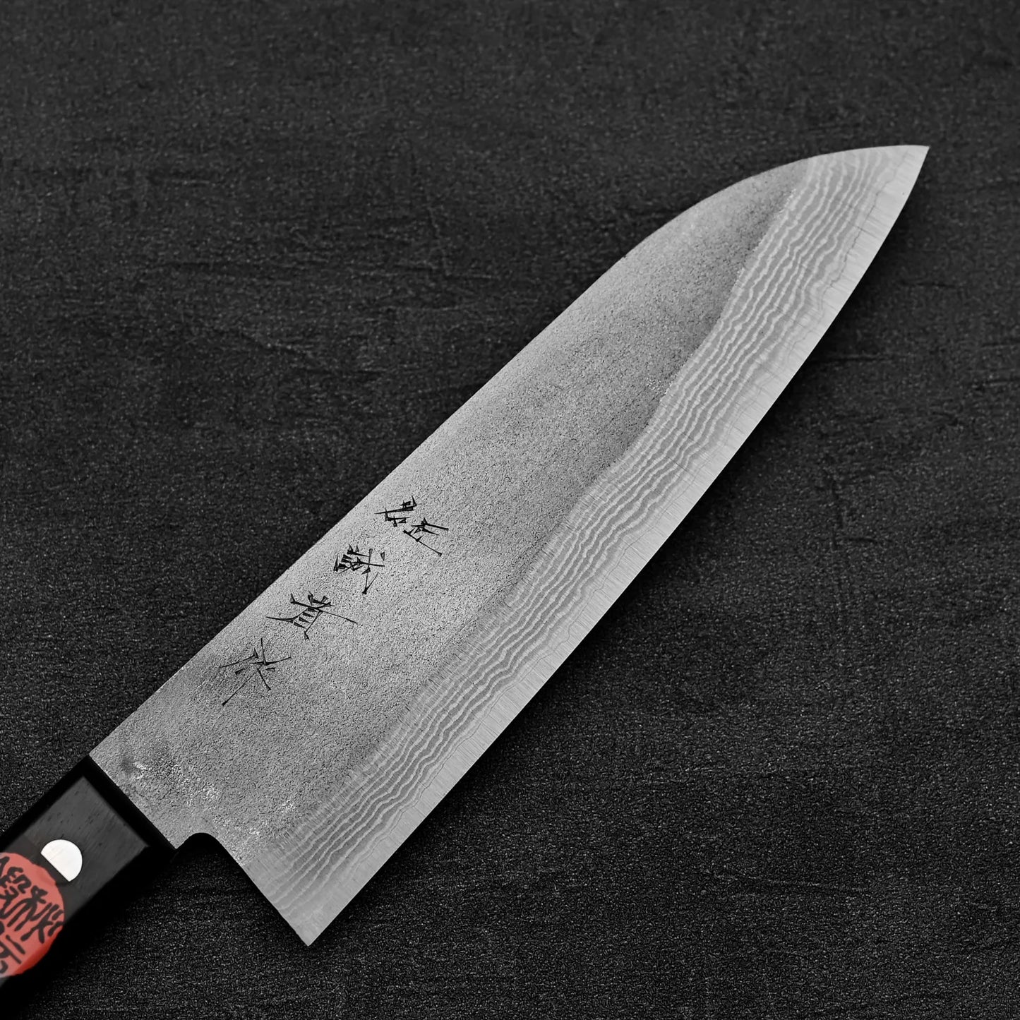 A Shigeki Tanaka nashiji damascus VG10 santoku kitchen knife with a black handle lies on a dark textured surface, featuring Japanese characters engraved near the handle.