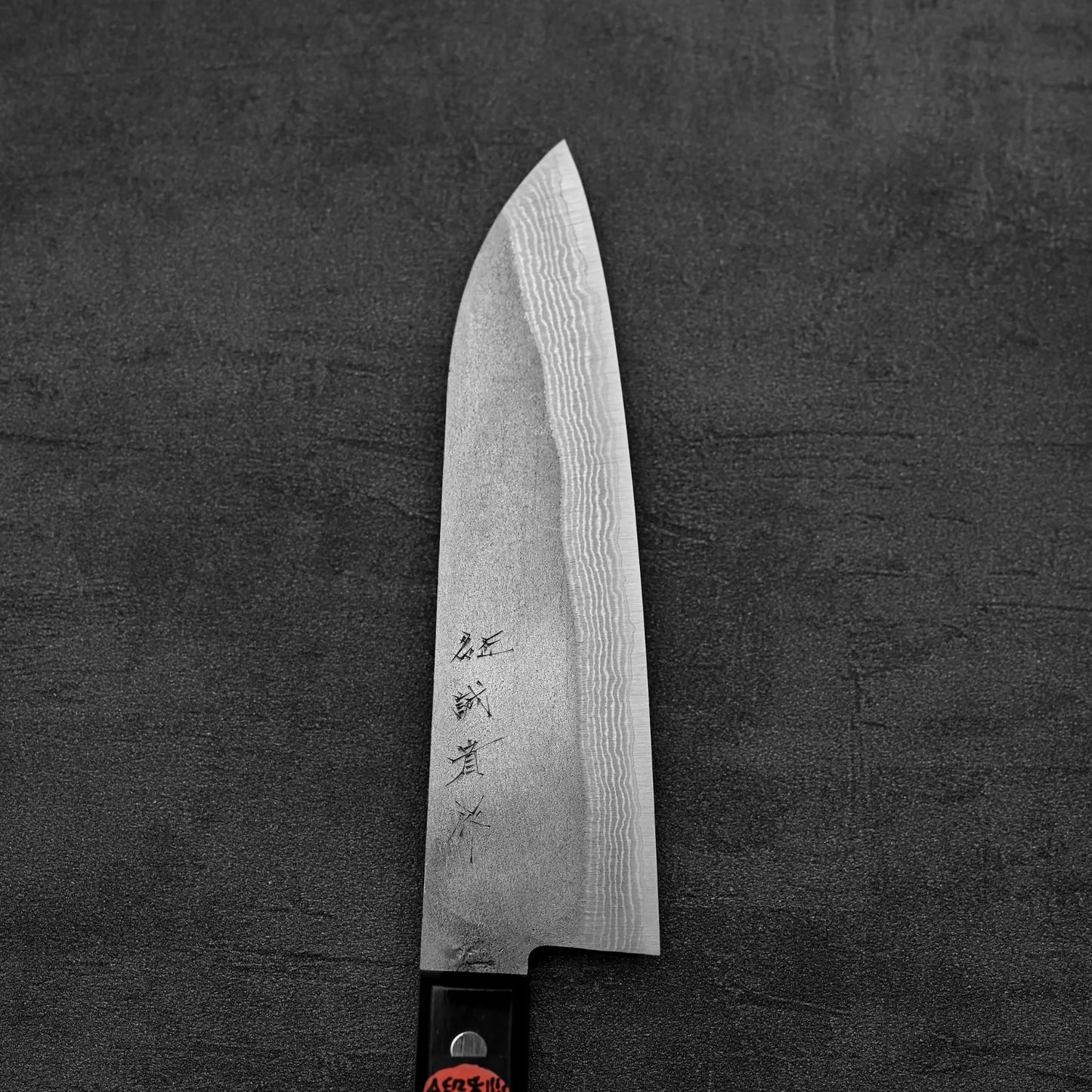 A Shigeki Tanaka nashiji damascus VG10 santoku by Shigeki Tanaka, with a damascus-patterned VG10 steel blade and engraved kanji, rests on a dark textured surface; its handle appears in the bottom left corner.