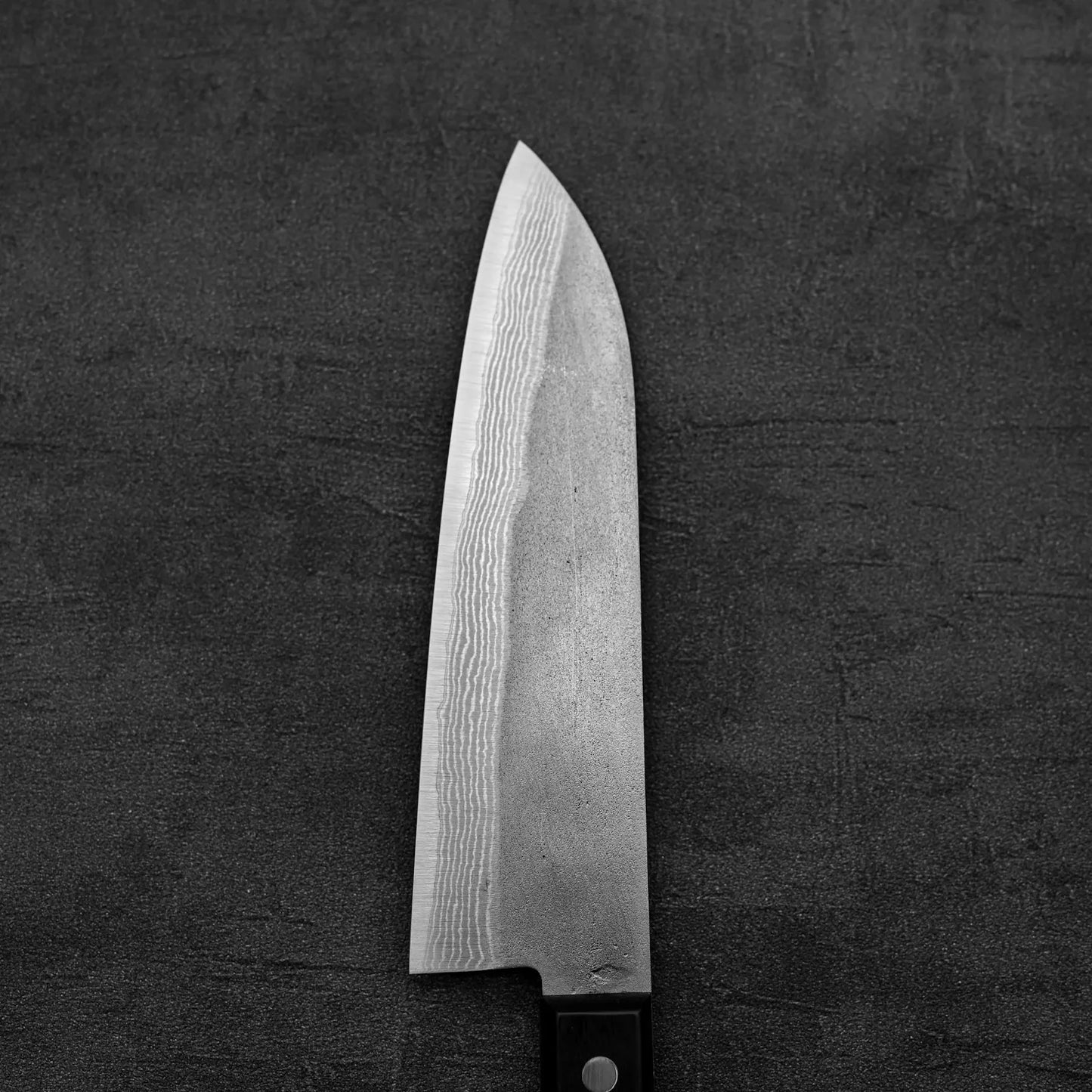 A close-up of the Shigeki Tanaka nashiji damascus VG10 santoku knife, featuring a distinctive damascus pattern on its VG10 steel blade by Shigeki Tanaka, shown on a dark textured surface.