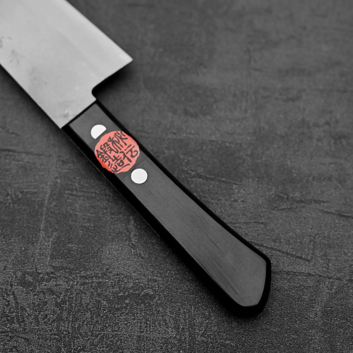A close-up of the Shigeki Tanaka nashiji damascus VG10 santoku kitchen knife, showing its black handle with two metal rivets and a red sticker with Japanese characters, placed on a dark textured surface.
