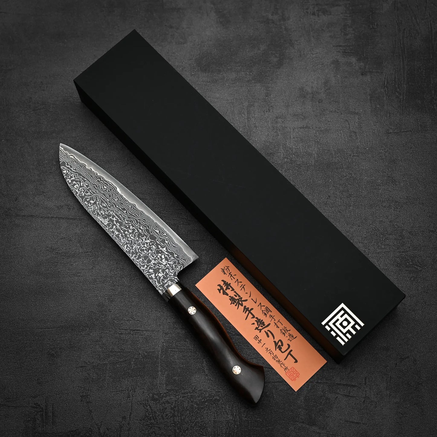 The Shigeki Tanaka R2 Damascus Santoku 170mm - Ironwood knife, with a patterned blade and dark wood handle, is placed beside a black box and an orange Japanese card on a dark textured surface.