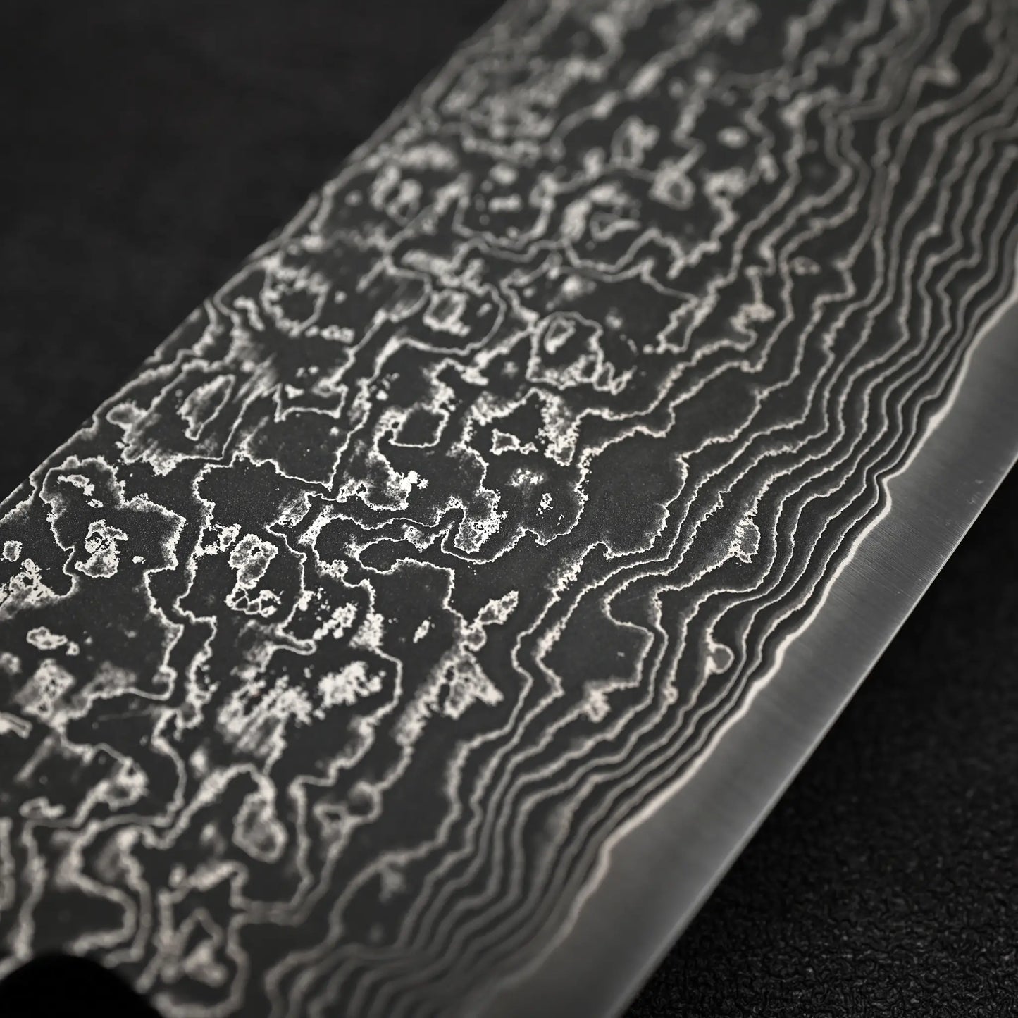 Close-up of the Shigeki Tanaka R2 Damascus Santoku 170mm - Ironwood, showing its intricate wavy blade patterns from layering and folding, set against a dark textured background.