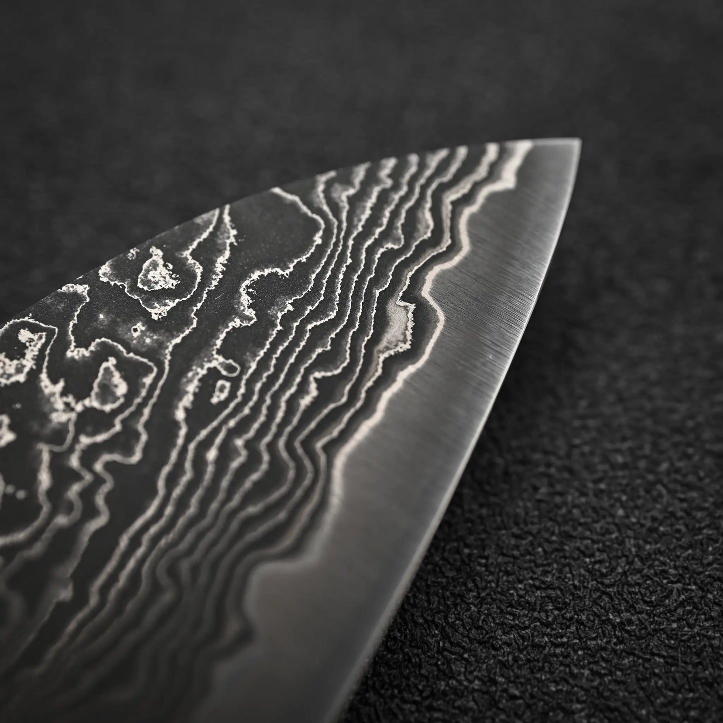 Close-up of the Shigeki Tanaka R2 Damascus Santoku 170mm - Ironwood blade, highlighting its intricate wavy patterns against a textured black background.