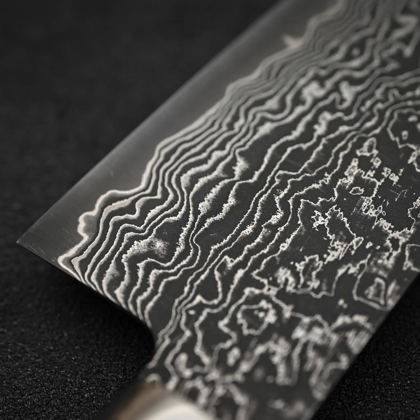Close-up of the Shigeki Tanaka R2 Damascus Santoku 170mm - Ironwood blade, highlighting its intricate wavy layered pattern, set against a dark textured background.