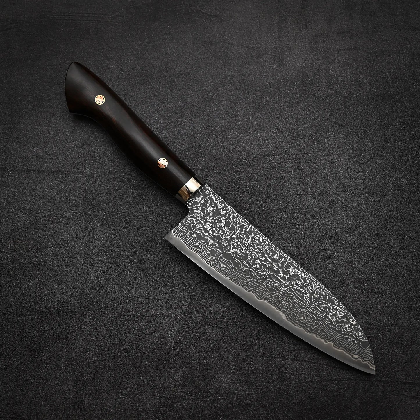 A Shigeki Tanaka R2 damascus santoku 170mm with an ironwood handle rests on a black, textured surface.