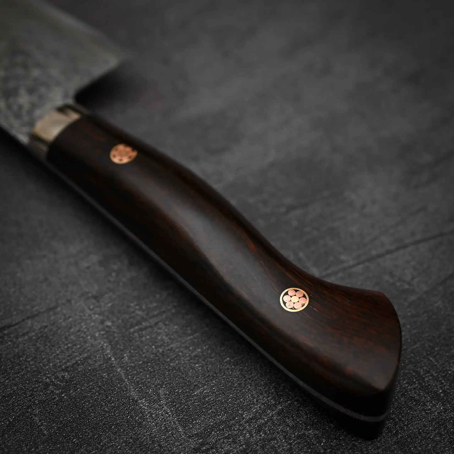 A close-up of the Shigeki Tanaka R2 Damascus Santoku 170mm’s ironwood handle with two metal rivets, set on a dark textured surface—a striking match for the renowned brands premium kitchen knife.