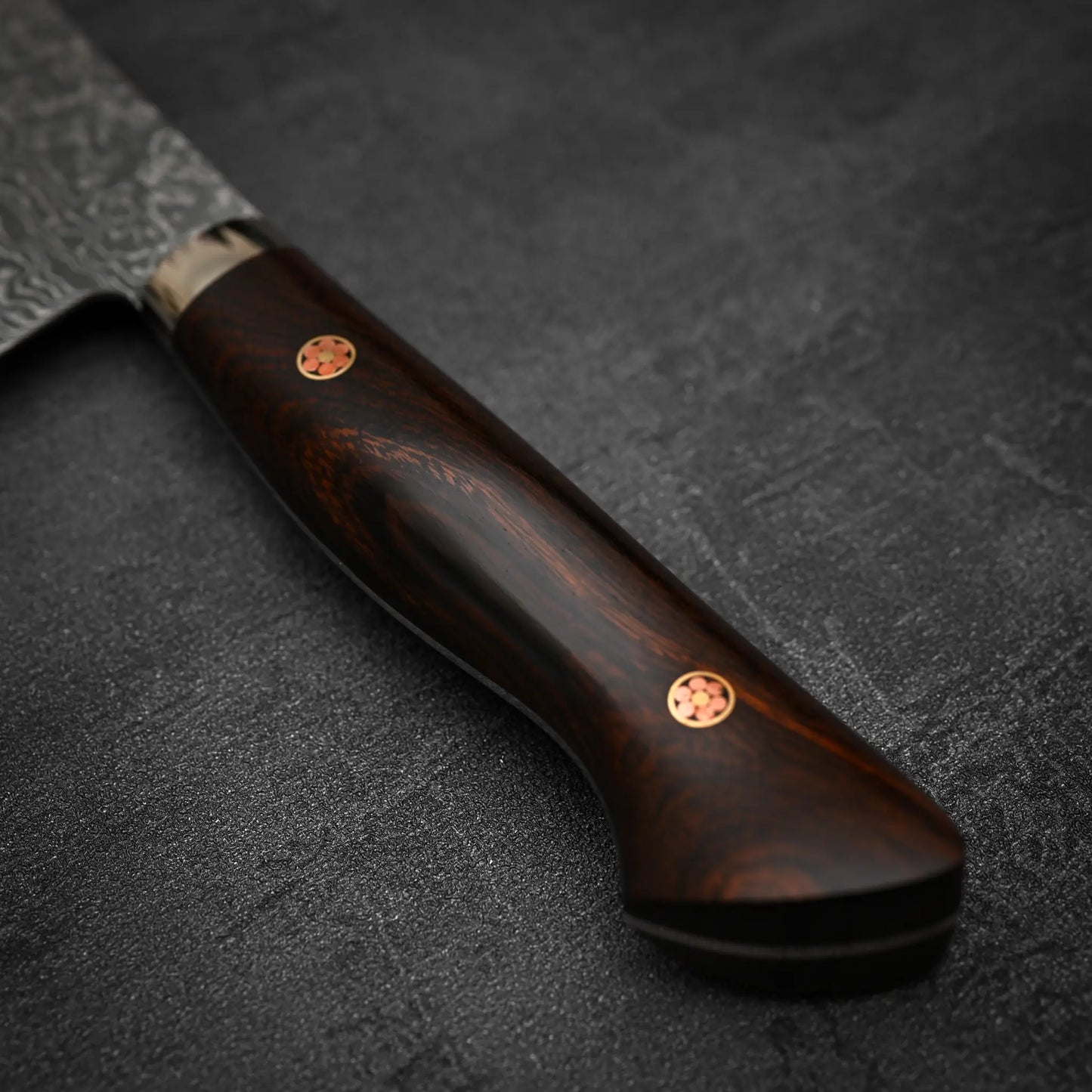 Close-up of the Shigeki Tanaka R2 Damascus Santoku 170mm - ironwood: its dark wooden handle with two brass pins rests on a textured surface, while the SG2 steel blade by Shigeki Tanaka appears at the top left.