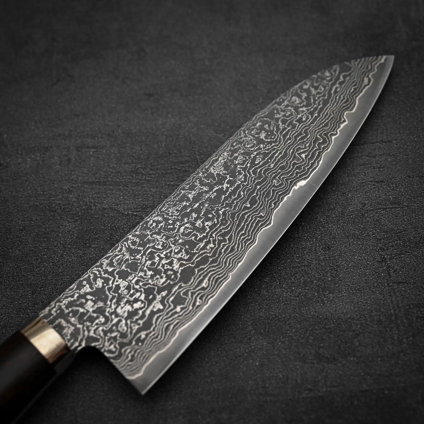 A close-up of the Shigeki Tanaka R2 damascus santoku 170mm - ironwood shows its dark handle and intricate wavy patterns on the SG2 steel blade, set against a dark, textured background.