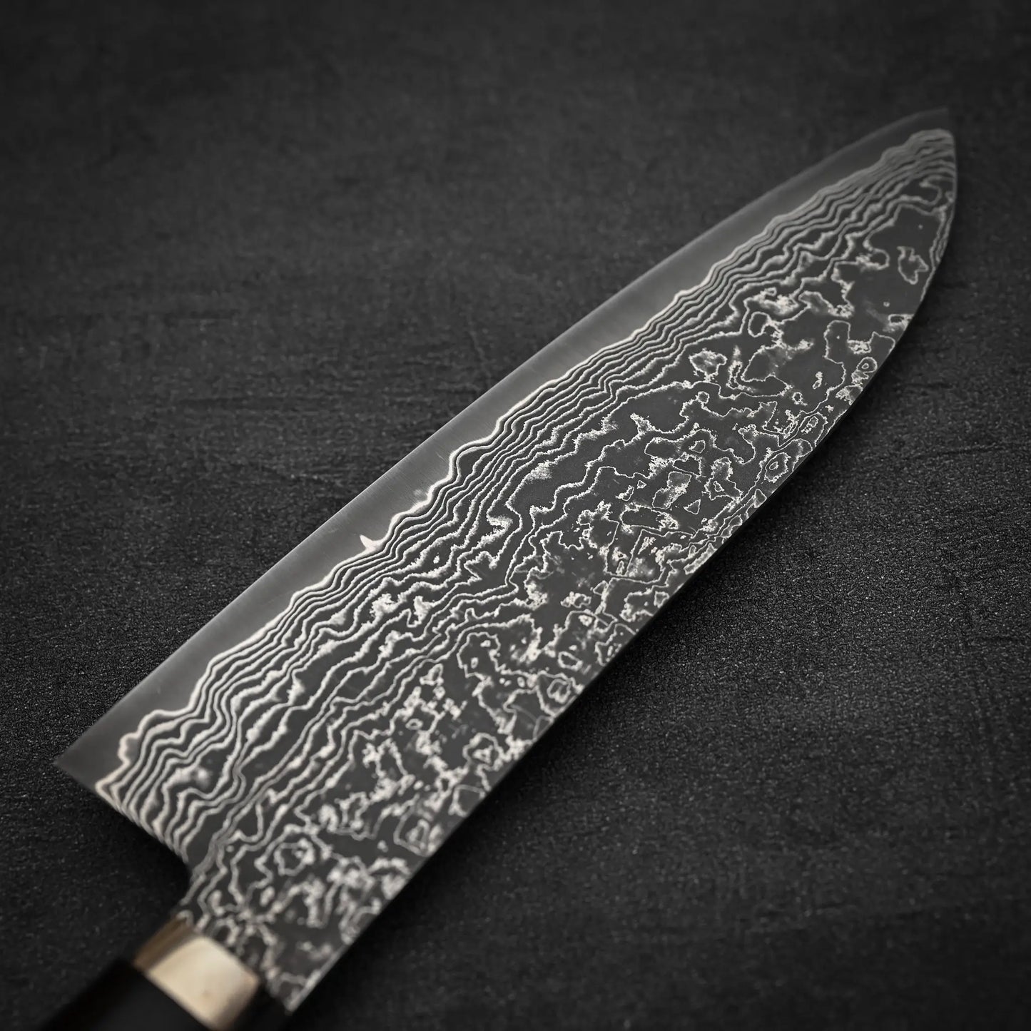 A close-up of the Shigeki Tanaka R2 Damascus Santoku 170mm - ironwood blade, displaying its intricate wavy patterns against a dark, textured background.