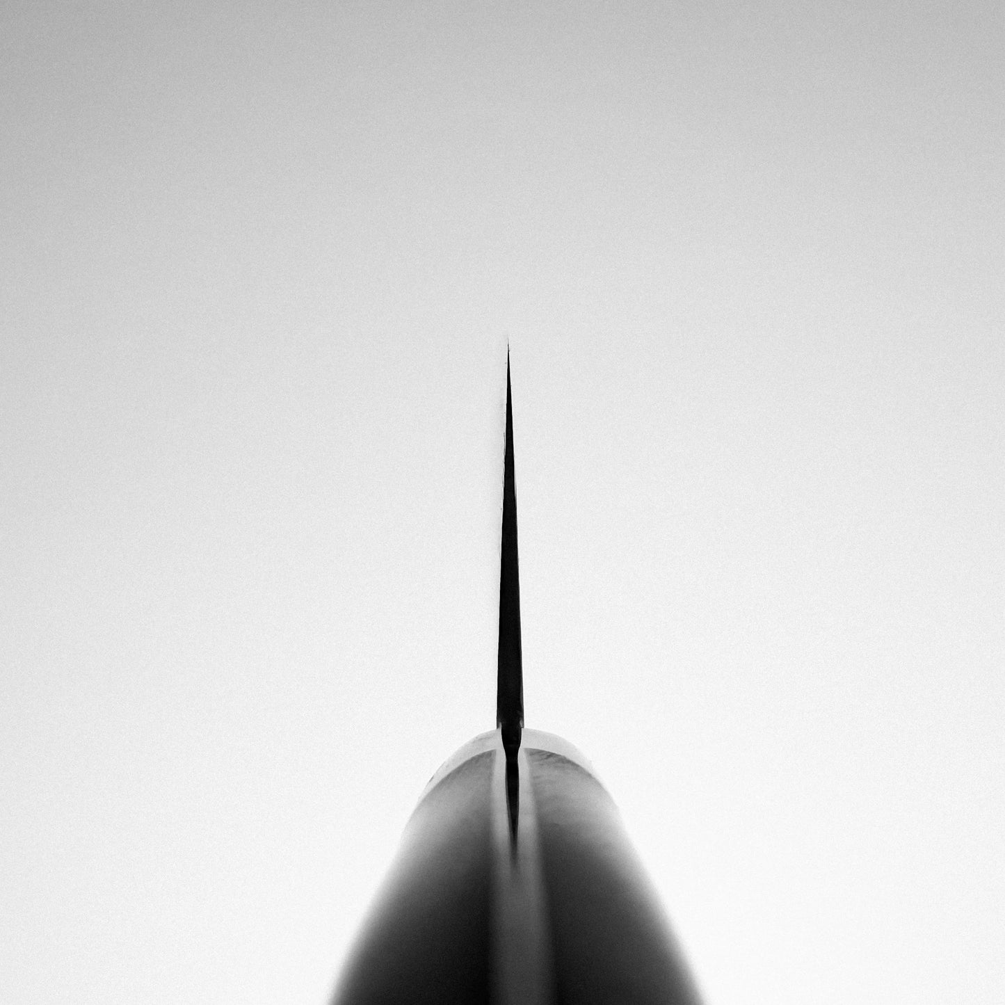 A black and white image shows the tip of the Shigeki Tanaka R2 Damascus Santoku 170mm - Ironwood, viewed from below with a symmetrical perspective against a blank sky. The sharply tapered blade highlights its refined edge.