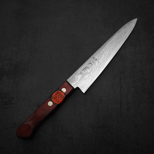 The Shigeki Tanaka VG10 damascus 150mm petty knife features a patterned silver blade with engraved symbols and a dark wooden handle with brass rivets and orange label, displayed on a black textured surface.