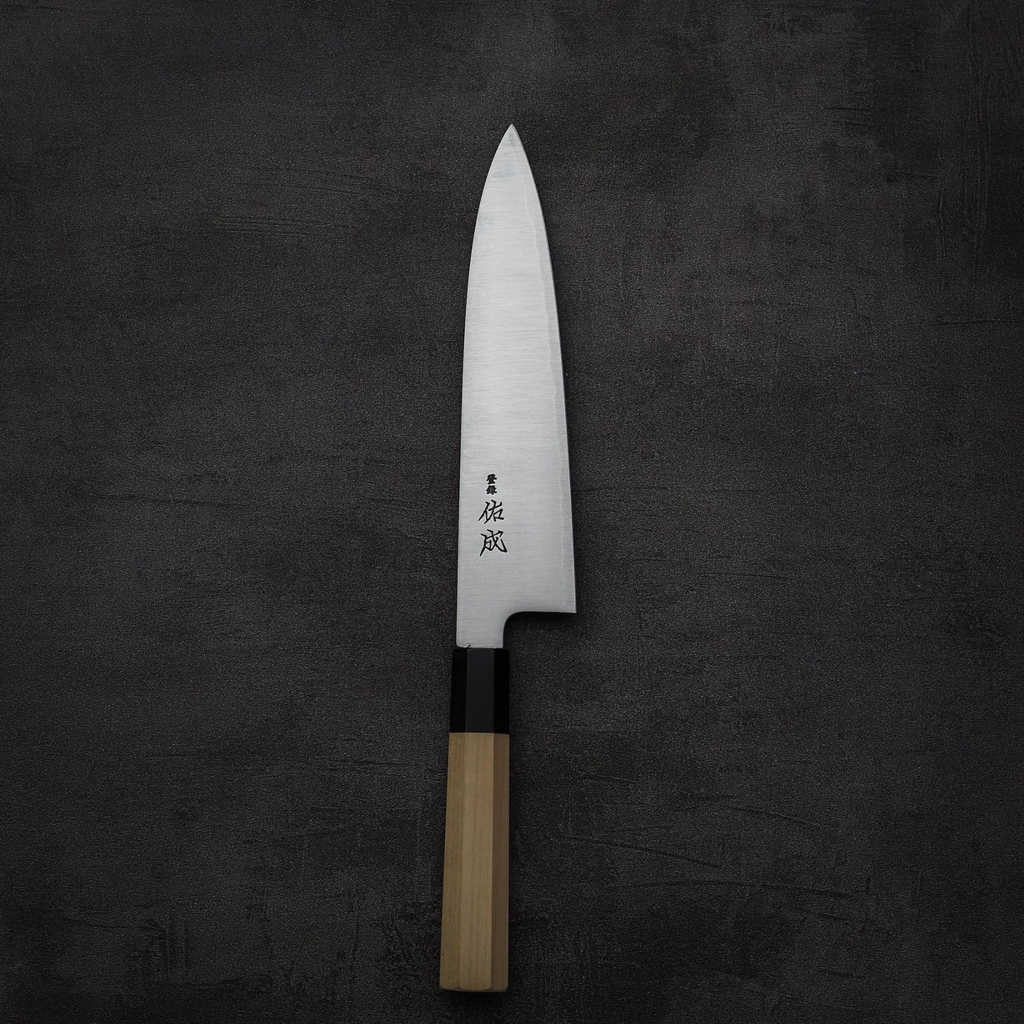 A Sukenari SG2 210mm gyuto chef knife by Sukenari, featuring a sharp SG2 steel blade and light wooden handle with magnolia saya, rests on a dark textured surface. Japanese characters are engraved near the handle.
