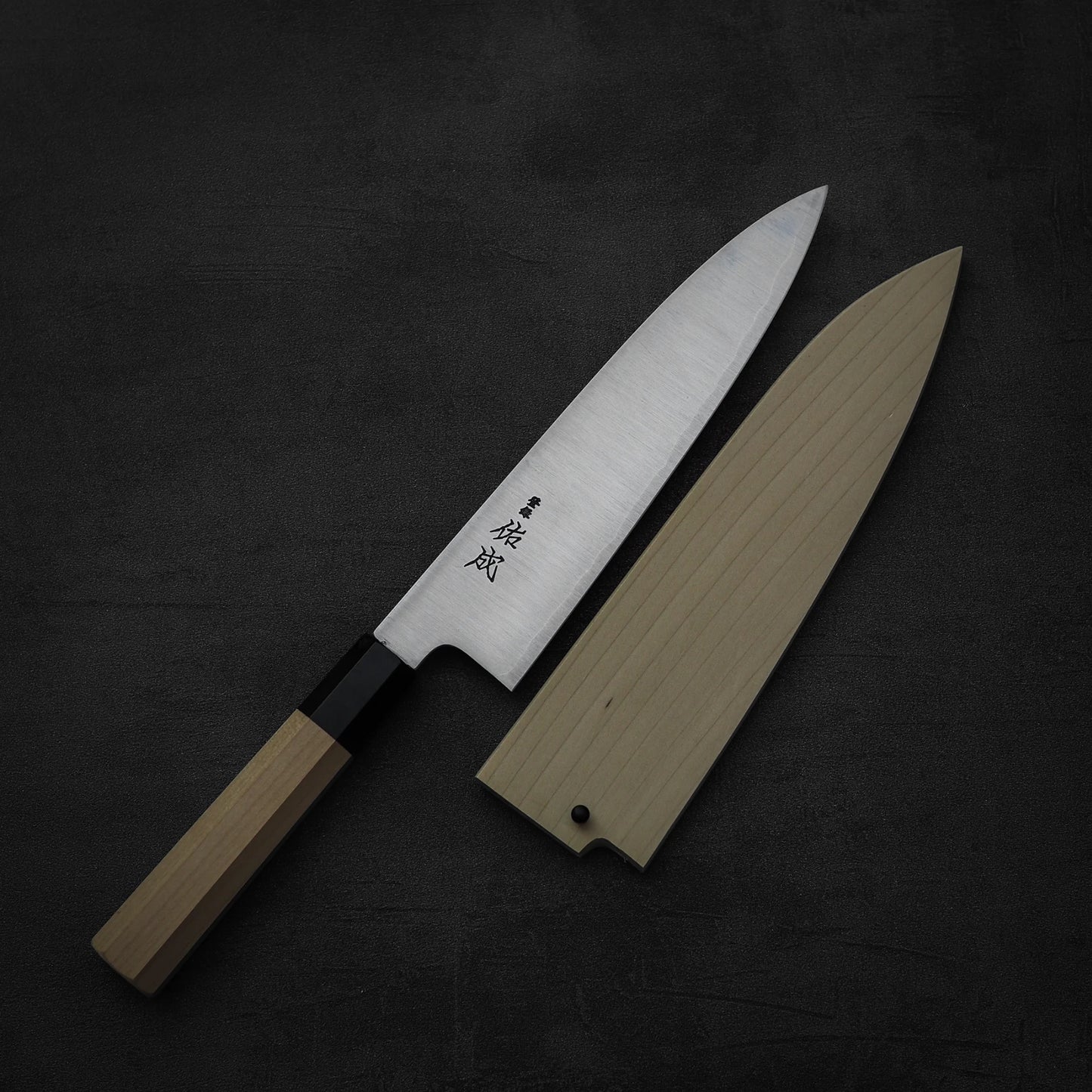 A Sukenari SG2 210mm gyuto chef knife with a wooden handle and engraved kanji, paired with a magnolia saya, is displayed on a dark textured background.