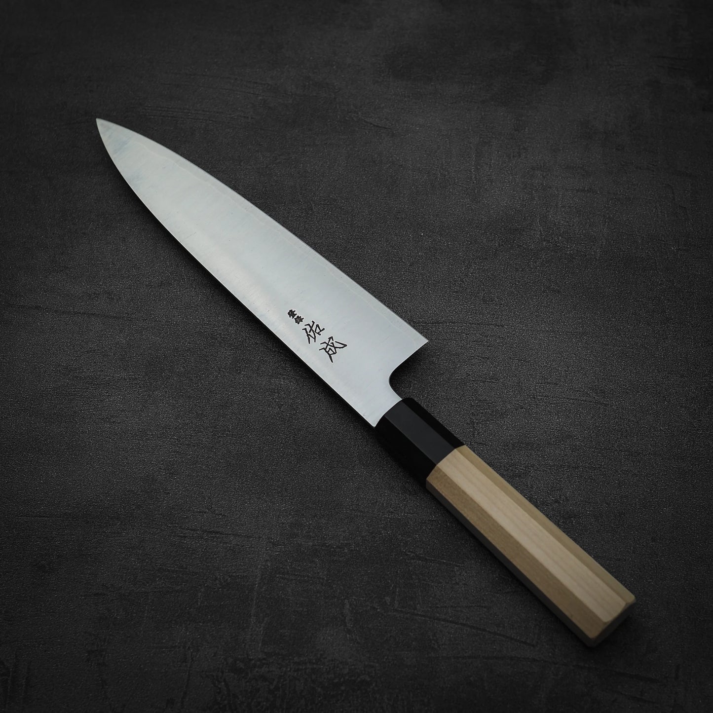 A Sukenari SG2 210mm gyuto with a light wooden handle and magnolia saya rests on a dark, textured surface. The blade displays Japanese characters near the handle, showcasing Sukenaris renowned craftsmanship.