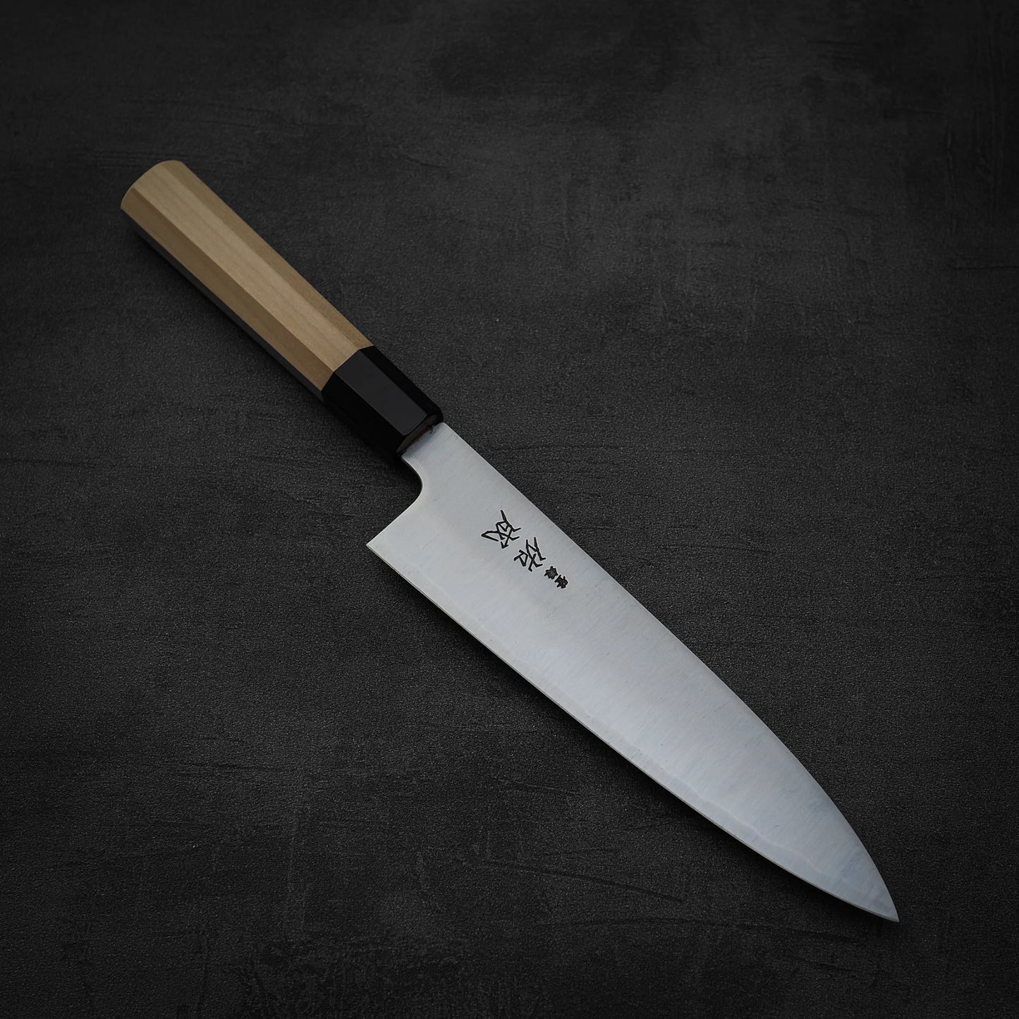 The Sukenari SG2 210mm gyuto, featuring a magnolia saya, rests on a dark, textured surface.