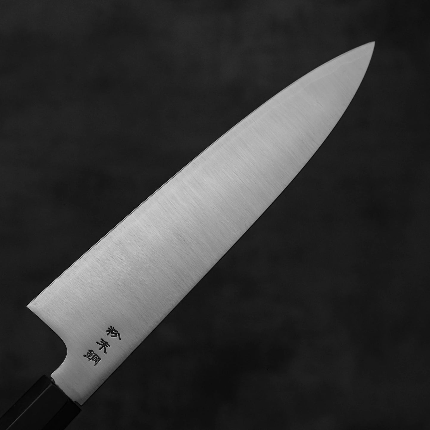 A close-up of the Sukenari SG2 210mm gyuto chef knife with a magnolia saya, featuring a black handle and engraved Japanese characters on the blade, set against a dark, blurred background.