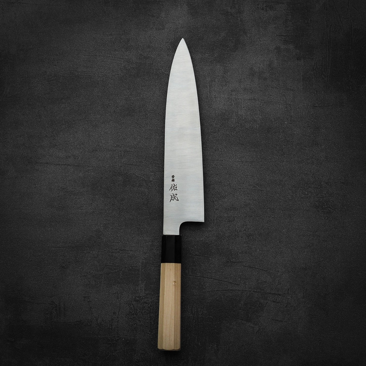 A Sukenari SG2 240mm gyuto from Sukenari, featuring a sharp blade and magnolia saya, rests on a dark textured surface. Japanese characters engraved on its side reflect its renowned edge retention.
