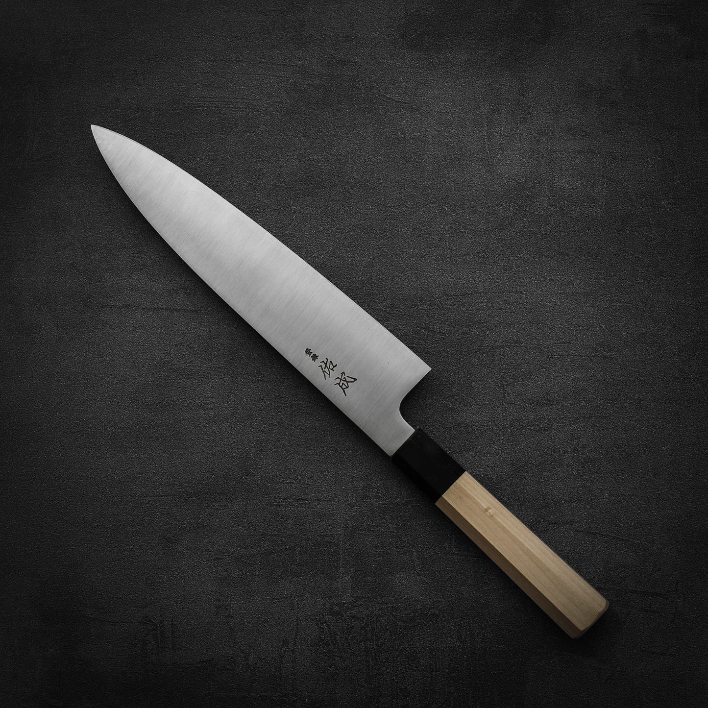 The Sukenari SG2 240mm gyuto, featuring a sharp steel blade and light wooden handle with magnolia saya, rests on a dark textured surface and is renowned for its long-lasting edge retention.