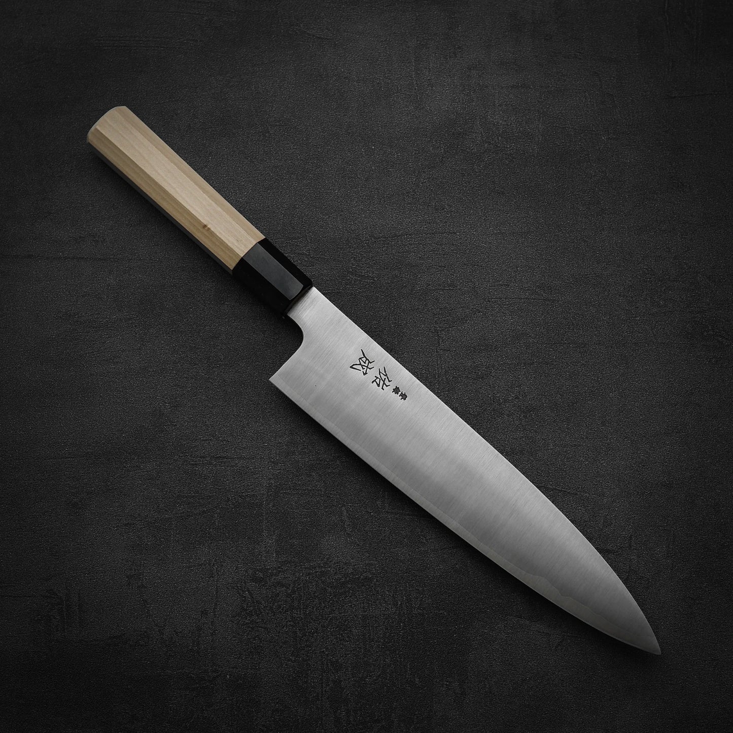 The Sukenari SG2 240mm gyuto, featuring a stainless steel blade and light wooden handle, displays Japanese engravings near its base. The knife, by Sukenari, is shown resting on a dark textured surface with its magnolia saya.