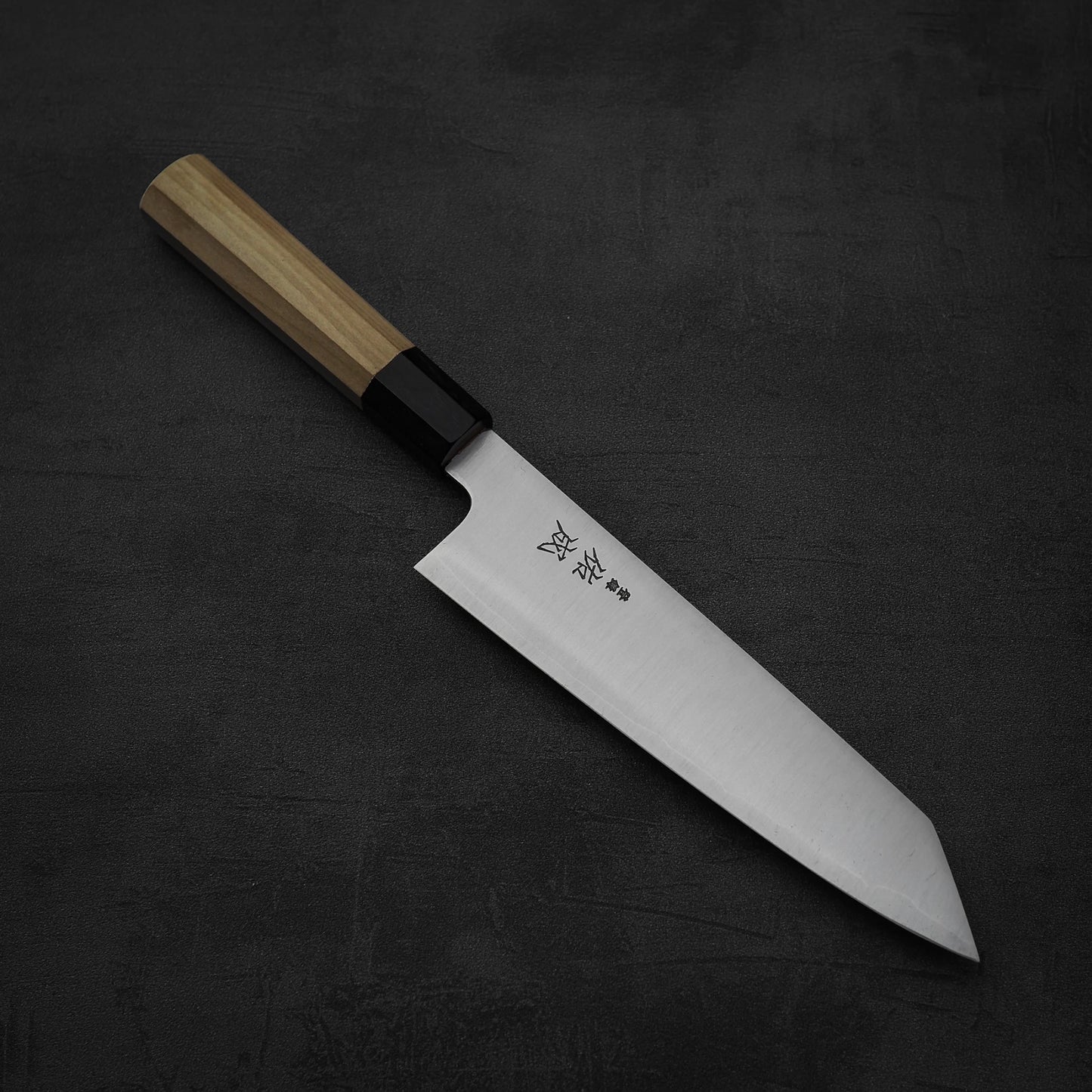 The Sukenari SG2 210mm kiritsuke gyuto chef knife, featuring a wooden handle and engraved Japanese blade, rests on a dark, textured surface. It comes with a magnolia saya and is expertly crafted by Sukenari.