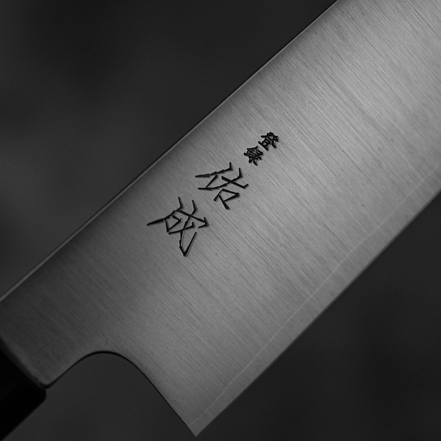 Close-up of the Sukenari SG2 240mm kiritsuke gyuto (with magnolia saya) chef knife blade by Sukenari, featuring Japanese engravings, set against a dark blurred background.