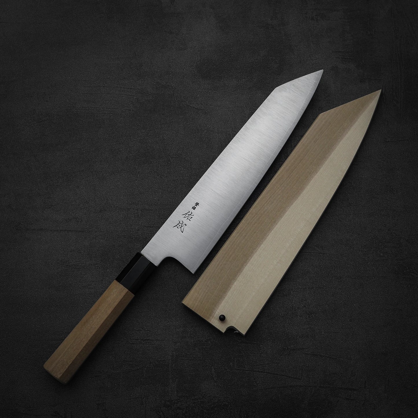 The Sukenari SG2 240mm kiritsuke gyuto by Sukenari, with a wooden handle and silver blade, rests beside its magnolia saya on a dark textured surface.