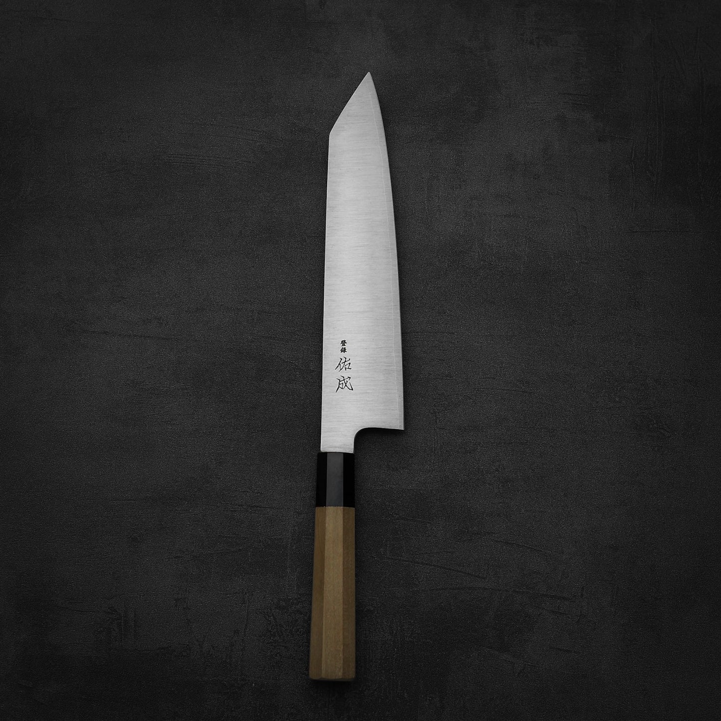 A Sukenari SG2 240mm kiritsuke gyuto with a magnolia saya and wide steel blade, featuring Japanese characters near the handle, rests on a dark, textured surface.