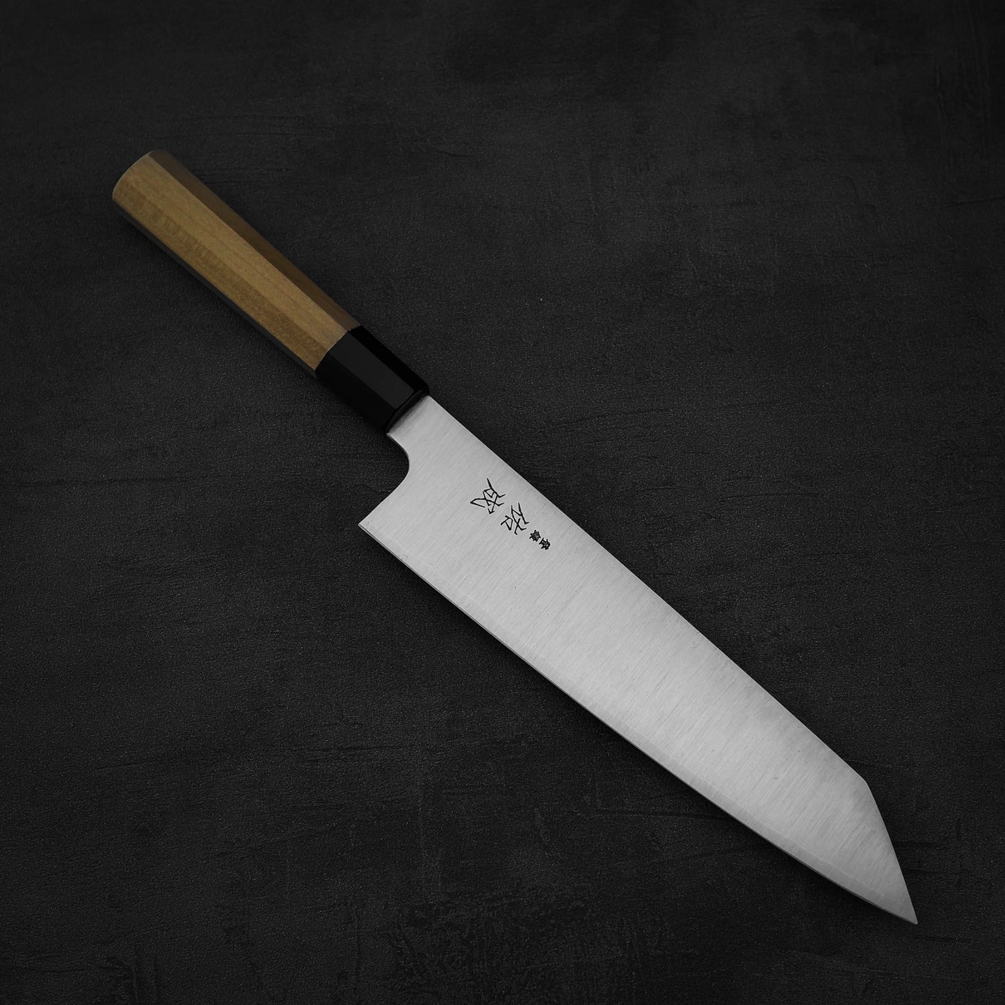 A Sukenari SG2 240mm kiritsuke gyuto by Sukenari, featuring a wooden handle and sharp stainless steel blade engraved with Japanese characters, rests on a dark textured surface. Comes with a magnolia saya.