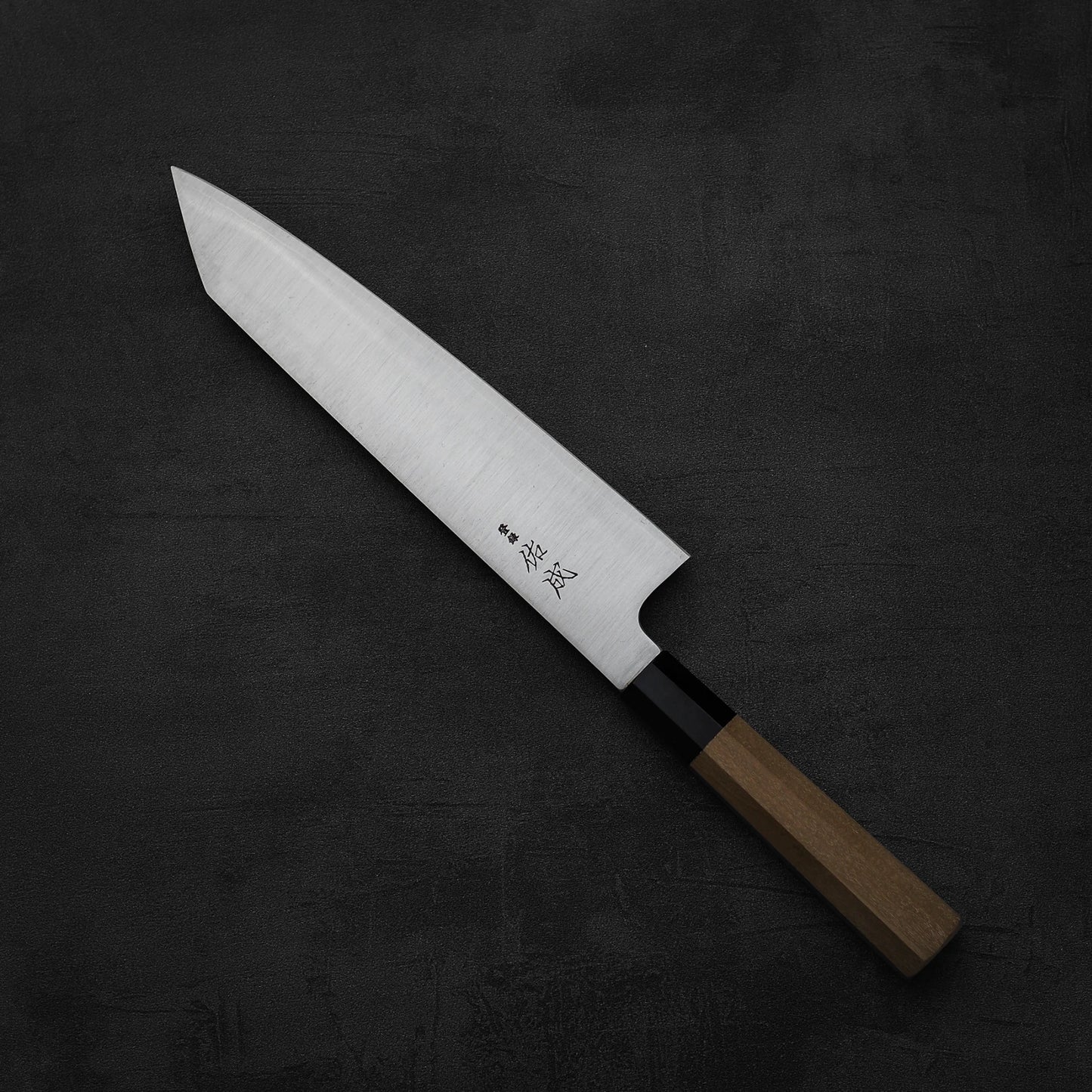 The Sukenari SG2 240mm kiritsuke gyuto chef knife, featuring a wooden handle and magnolia saya, rests on a dark textured background. Elegant Japanese engravings decorate the blade near the handle.