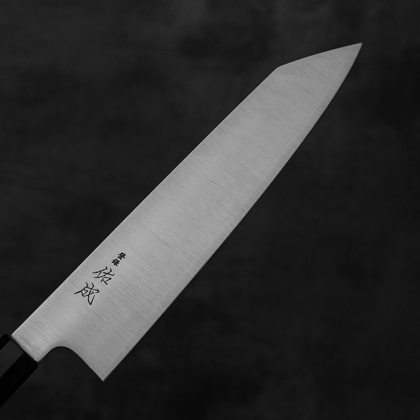 The Sukenari SG2 240mm kiritsuke gyuto chef knife by Sukenari features a sharp, pointed blade with Japanese characters near the handle and includes a magnolia saya, displayed on a dark gray background.