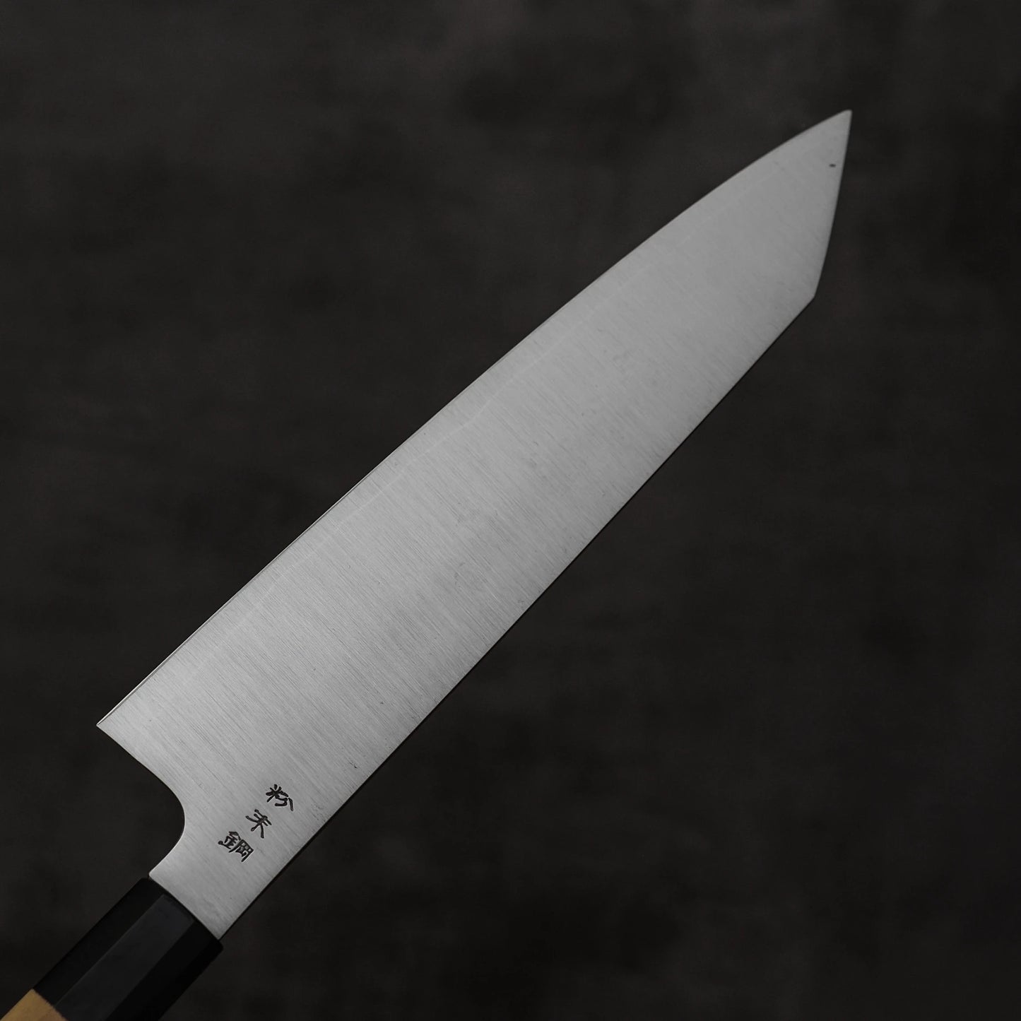 A close-up of a Sukenari SG2 240mm kiritsuke gyuto chef knife, featuring Japanese engravings near the handle, with a sharp pointed blade and magnolia saya, set against a dark, blurred background.