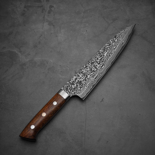 The Takeshi Saji black damascus SG2 gyuto ironwood 210mm, featuring a patterned stainless steel blade and ironwood handle with three metal rivets, rests on a dark, textured surface.