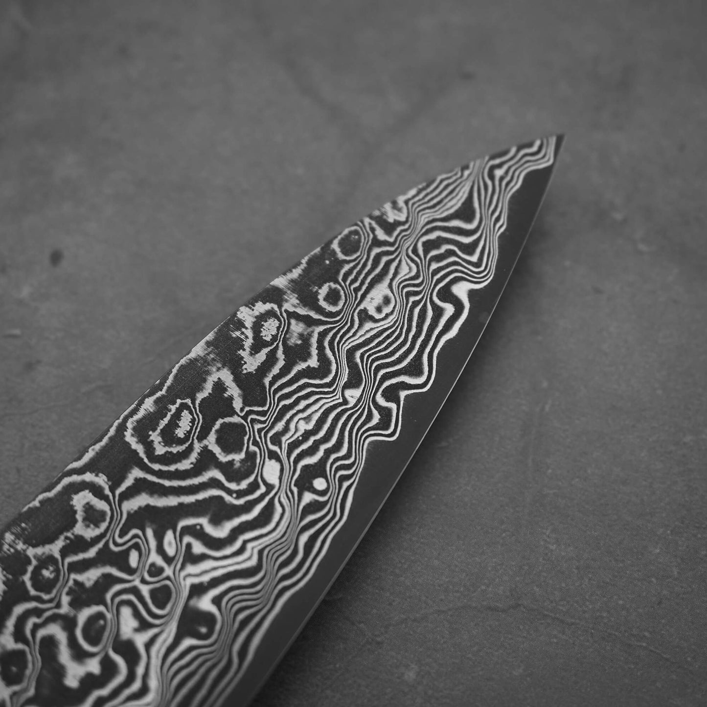 Close-up of the Takeshi Saji black damascus SG2 gyuto ironwood 240mm, featuring intricate wavy patterns on its SG2 steel blade, with detailed designs standing out against a plain background.
